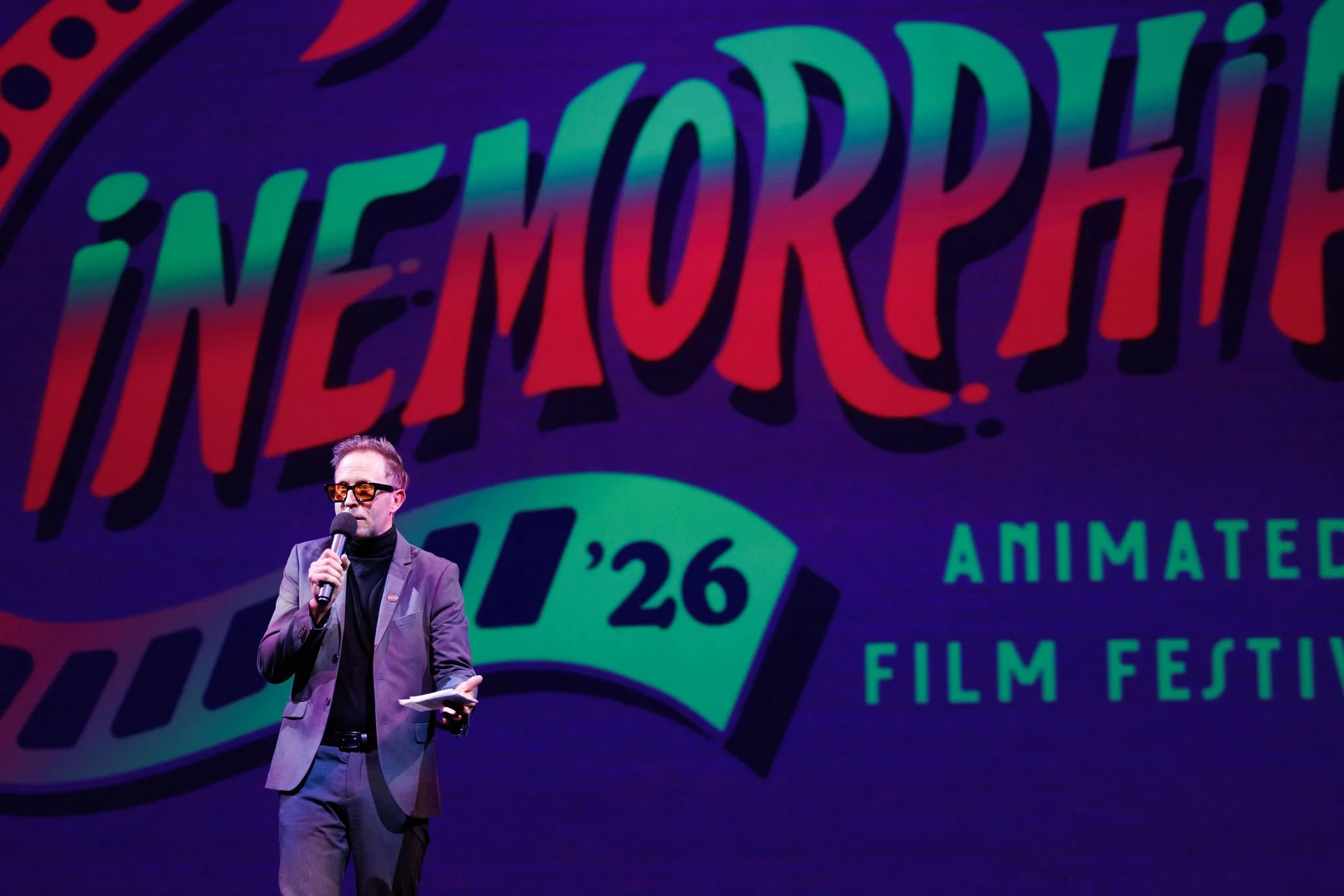 A man in a suit and sunglasses holding a microphone and paper, standing on stage inside Atlas9 in front of a colorful festival sign that reads 'Cinemorphia 26 Animated Film Festival'.