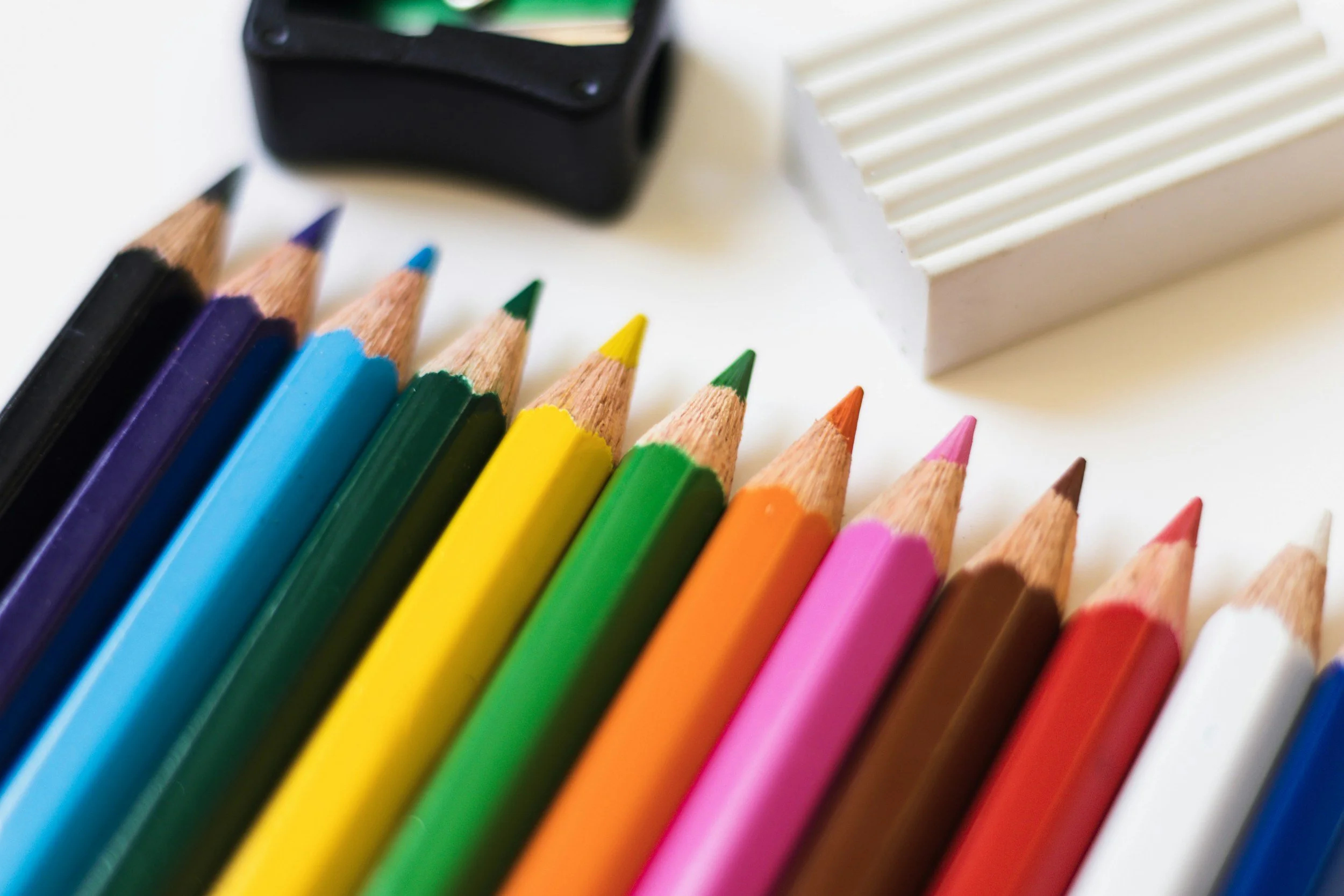 A set of colored pencils arranged in a row with the tips pointing upward, next to a white ribbed container and a black object.