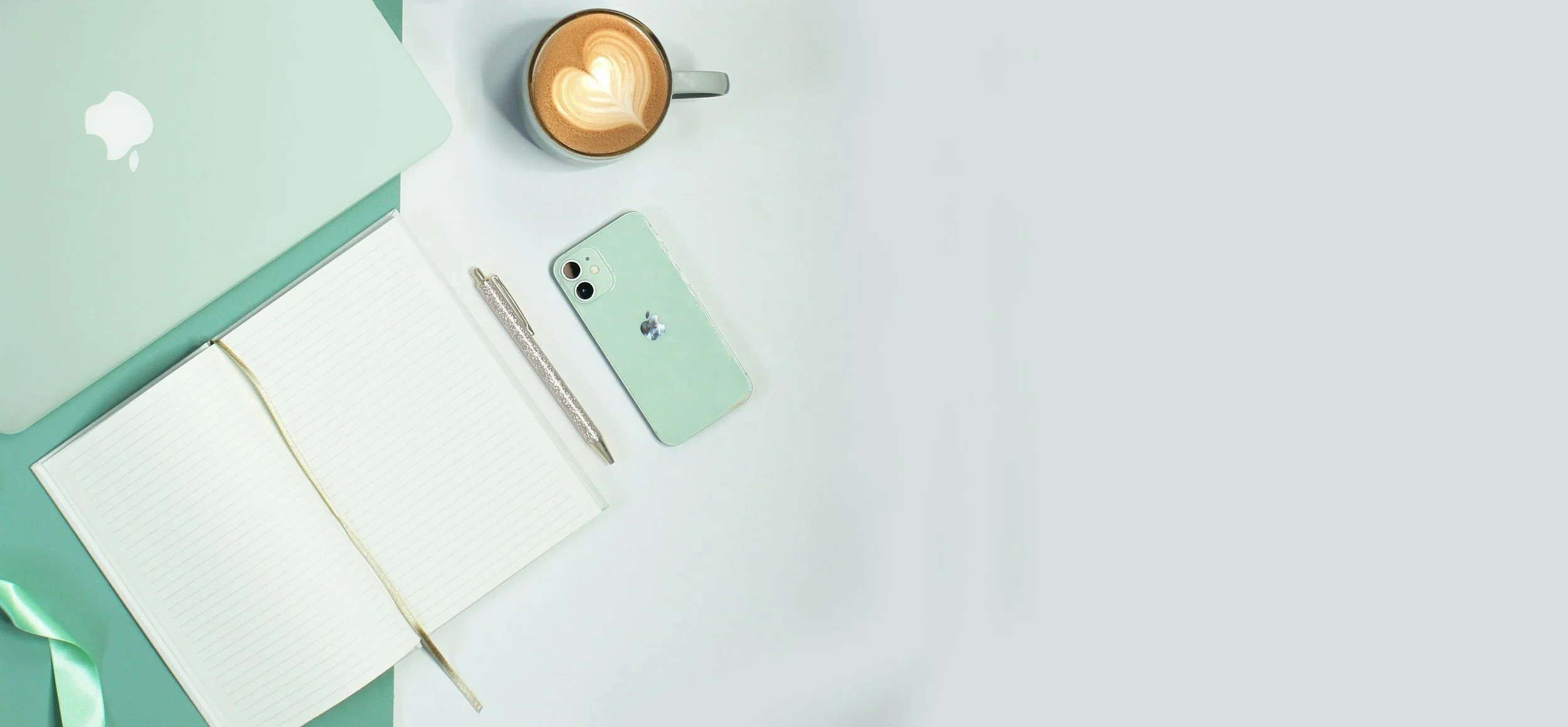 A flat lay of a workspace with a mint green MacBook, an open lined notebook, a sparkly pen, a mint green iPhone, and a cup of coffee with latte art on a white surface.