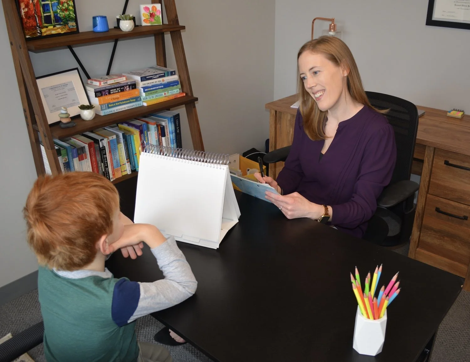 Dr. Christine Howard sits behind a black desk in her office, smiling and talking to a young boy with red hair, who is seated across from her. Dr. Howard and the boy work together to complete psychological testing.