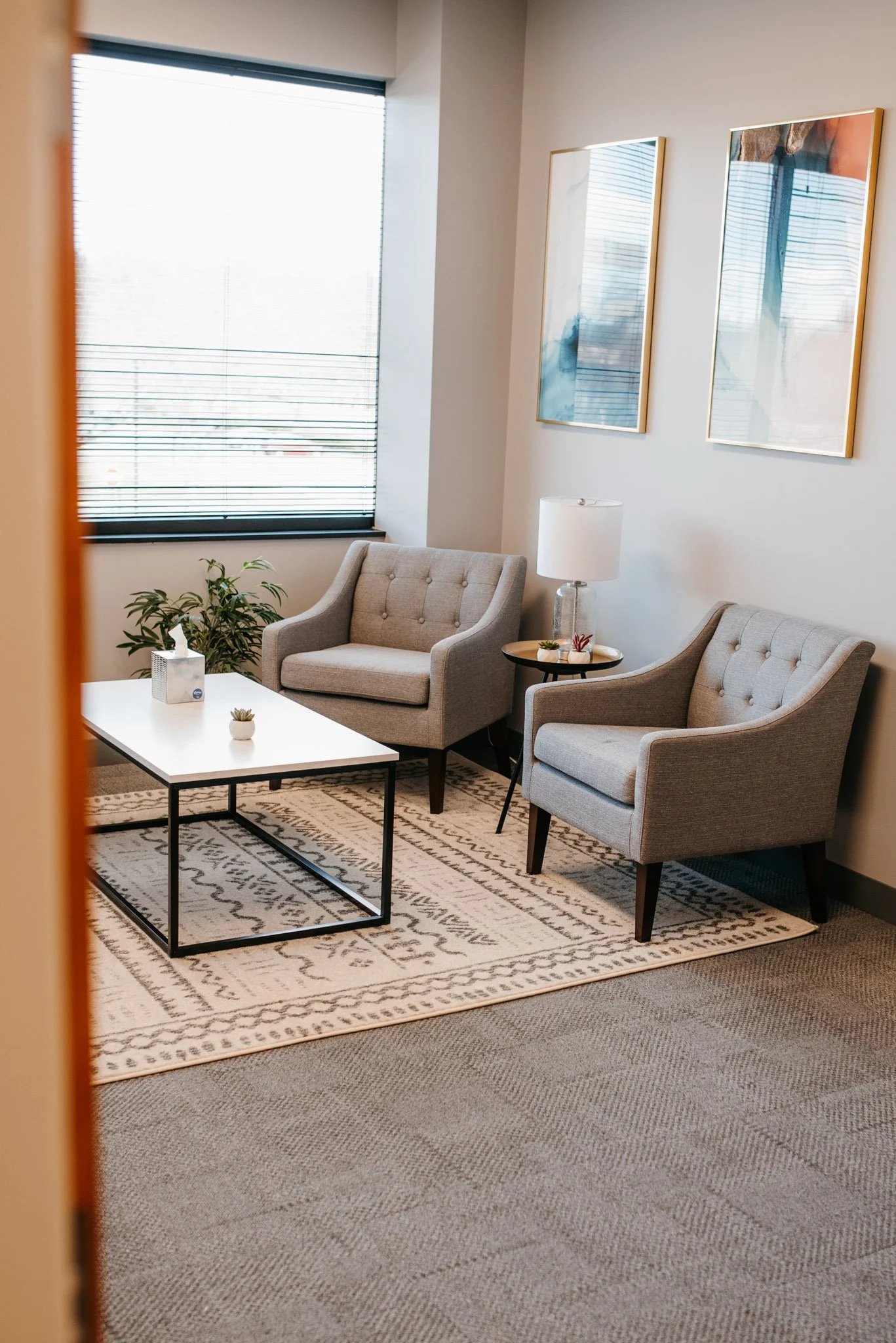 The inside of Dr. Christine Howard's therapy office. The room has a large window with horizontal blinds, a decorative rug, a gray carpeted floor, tufted chairs, and colorful artwork on the wall.