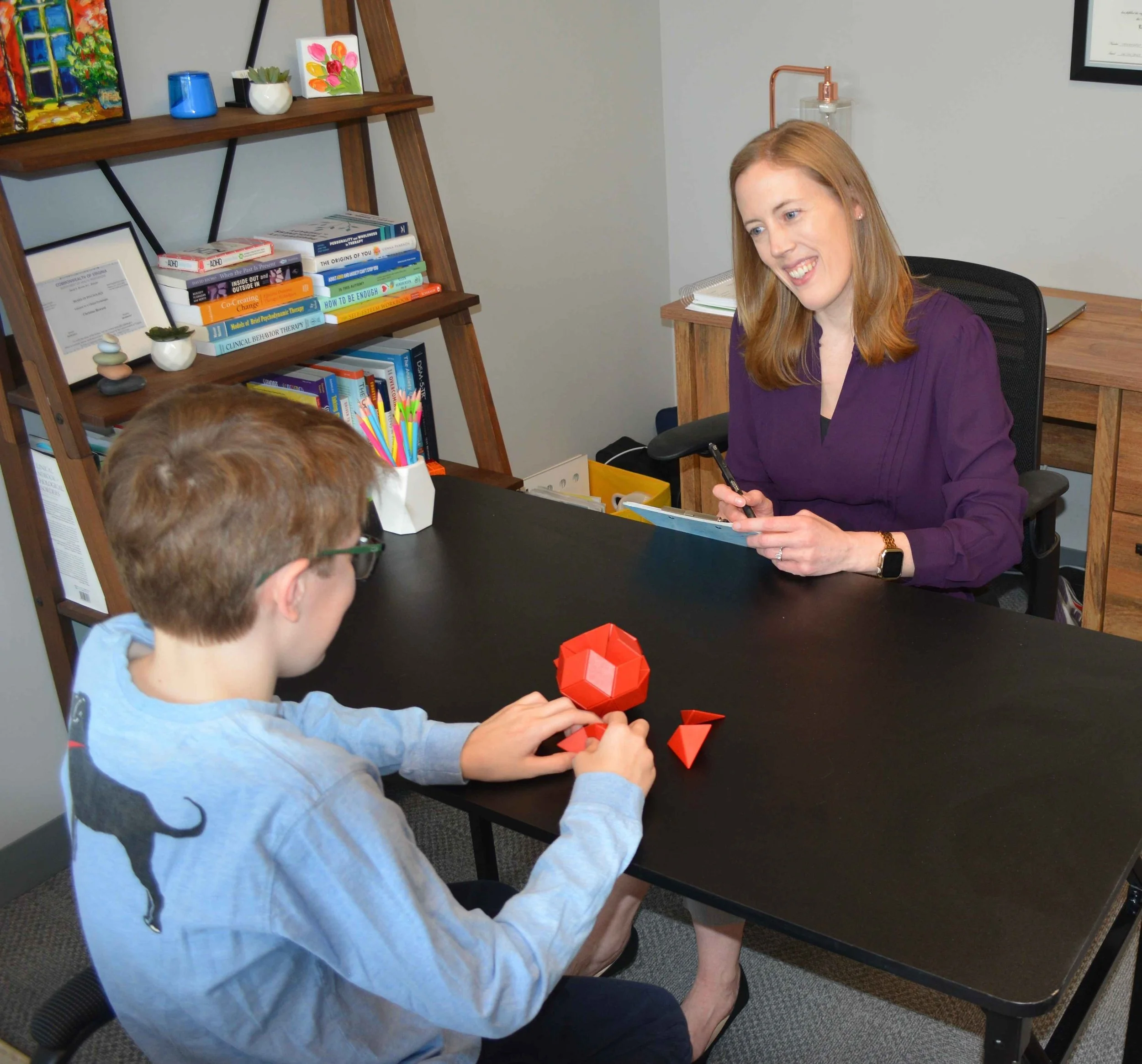 Dr. Christine Howard sits behind a black desk in her office, smiling and talking to a boy with brown hair, who is seated across from her. Dr. Howard and the boy work together to complete psychological testing.