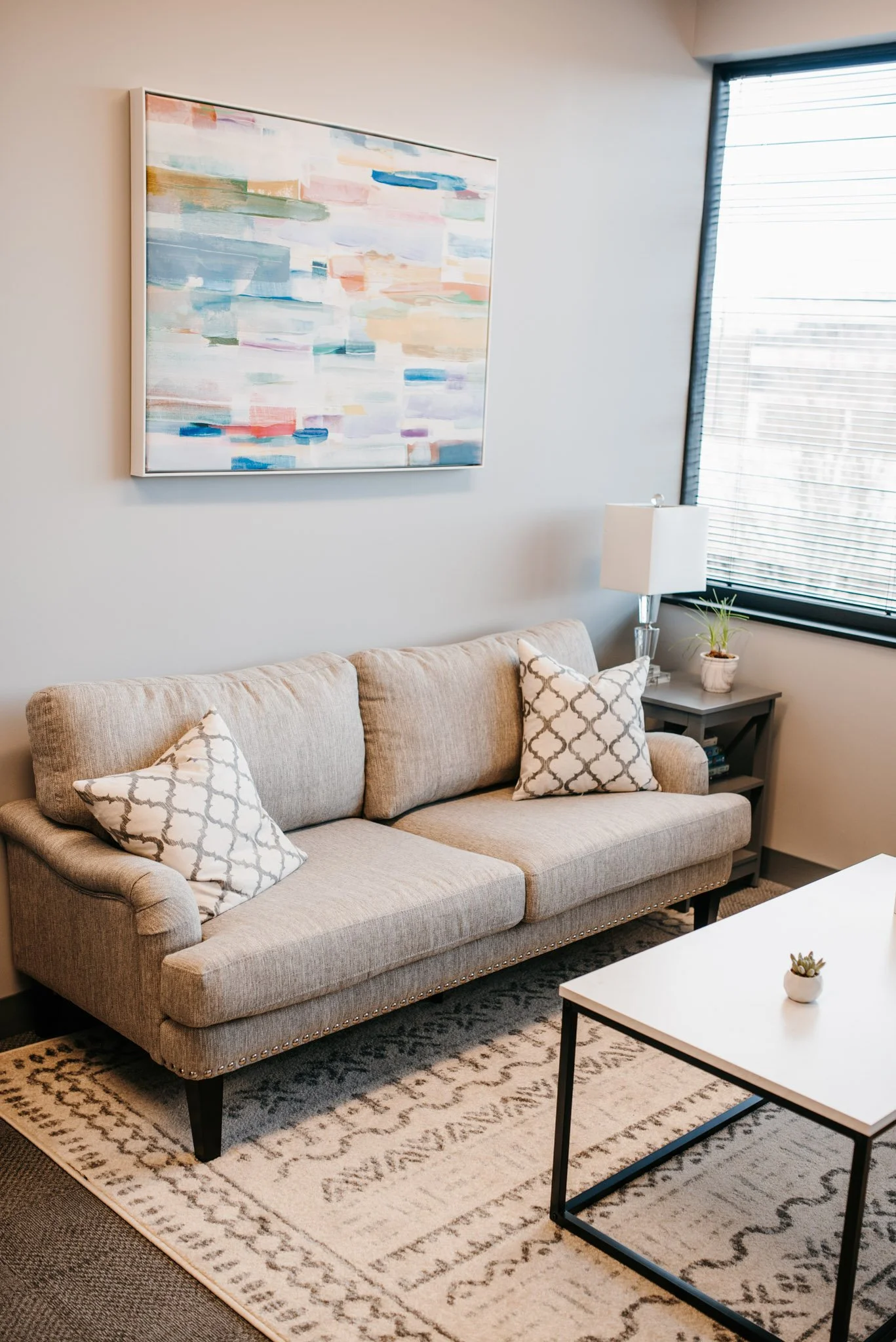 The inside of Dr. Christine Howard's therapy office. A beige sofa with pillows, a white coffee table with a small plant and tissues, a large abstract painting on the wall, a side table with a lamp and a small plant, and a window with blinds.