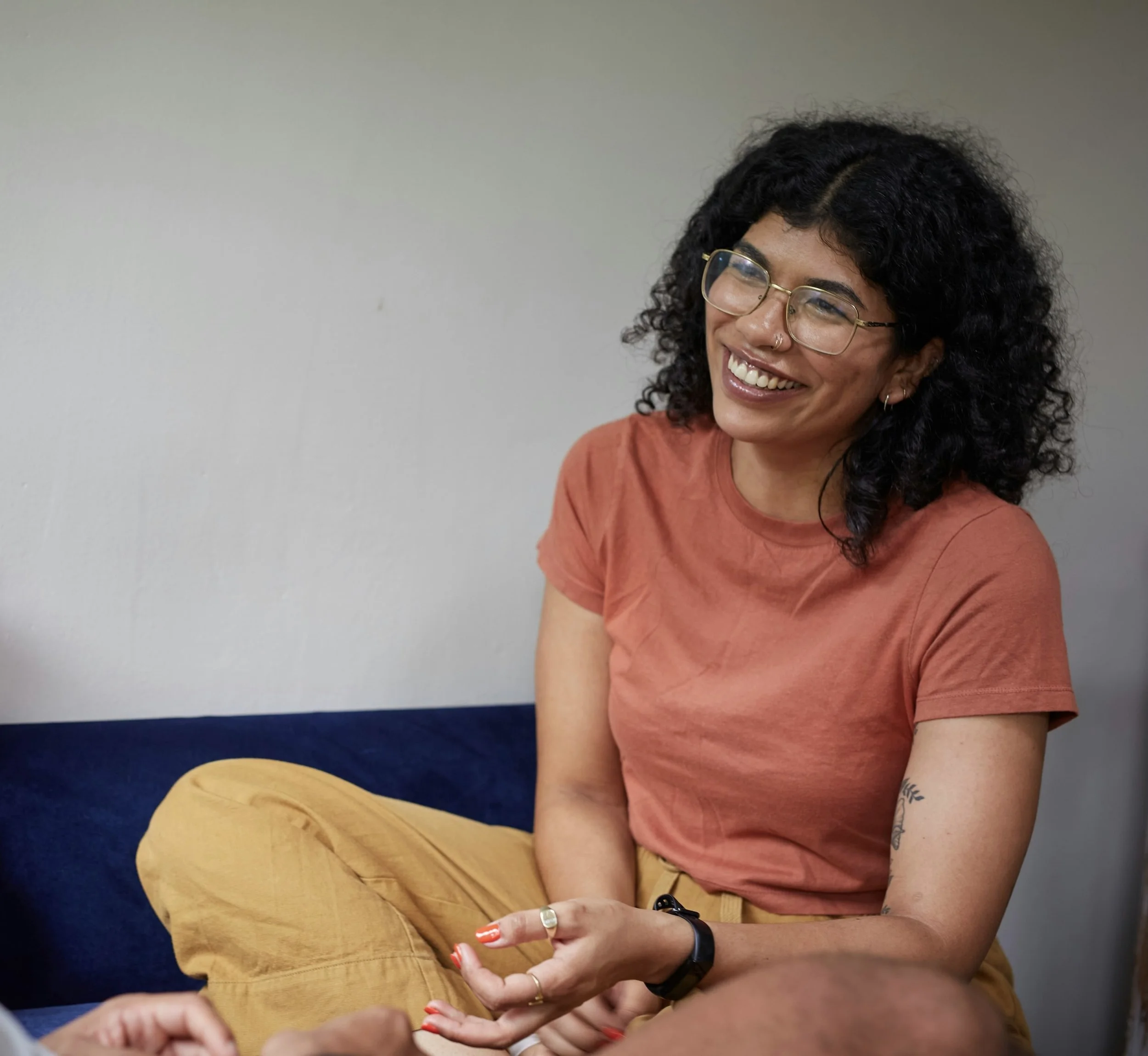 A woman with curly dark hair and glasses smiling while seated on a therapy couch, wearing a short-sleeved orange shirt, with a tattoo on her left arm, against a plain light-colored wall.