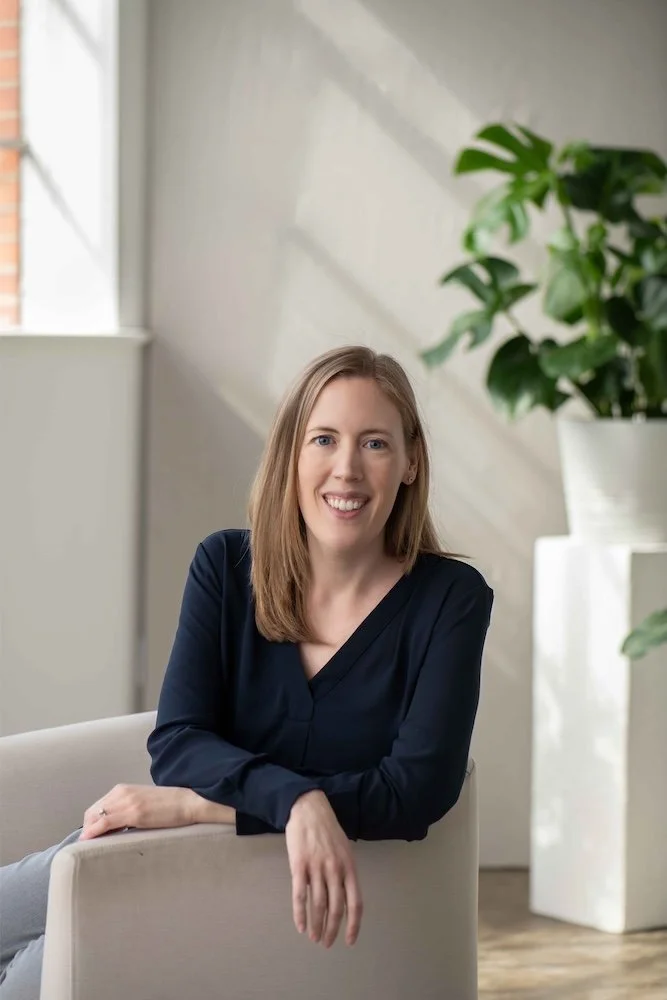 Dr. Christine Howard, a clinical psychologist who provides therapy and psychological testing, sitting on a beige armchair in a modern, minimalistic room with a large green plant in a white pot and a window with natural light.