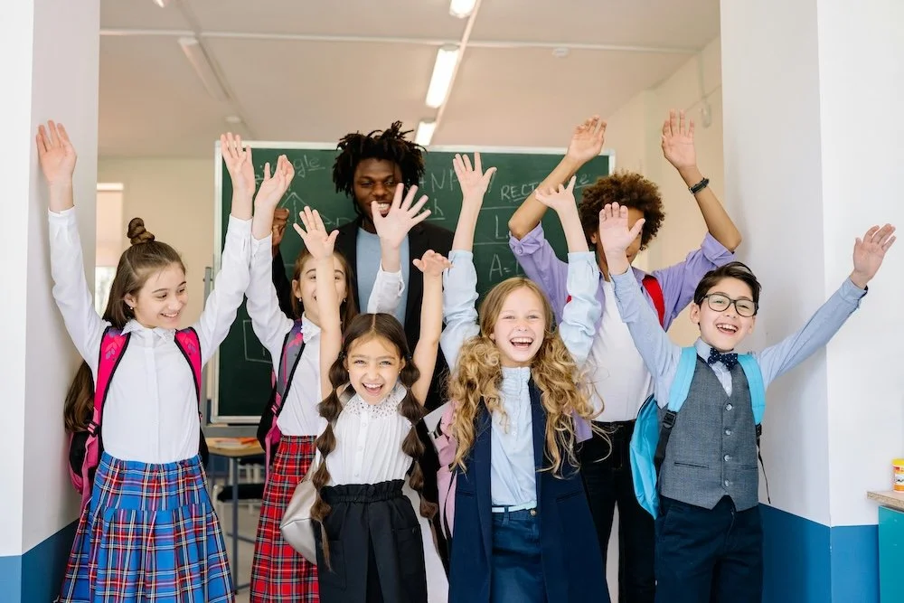 A group of children and a teacher raising their hands in a classroom. The children are wearing school uniforms, and the teacher is standing behind them with a blackboard with writing in the background.