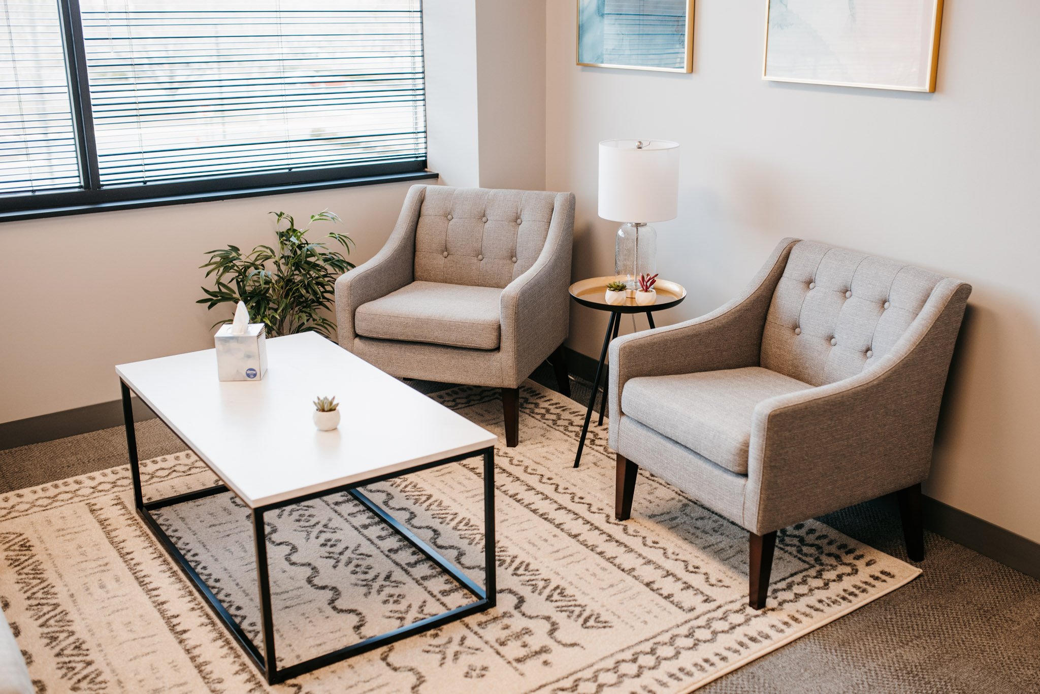 The inside of Dr. Christine Howard's office. There are two light gray tufted armchairs, a white coffee table, a clear table lamp, a plant, and framed artwork on the wall. There is a window with blinds.