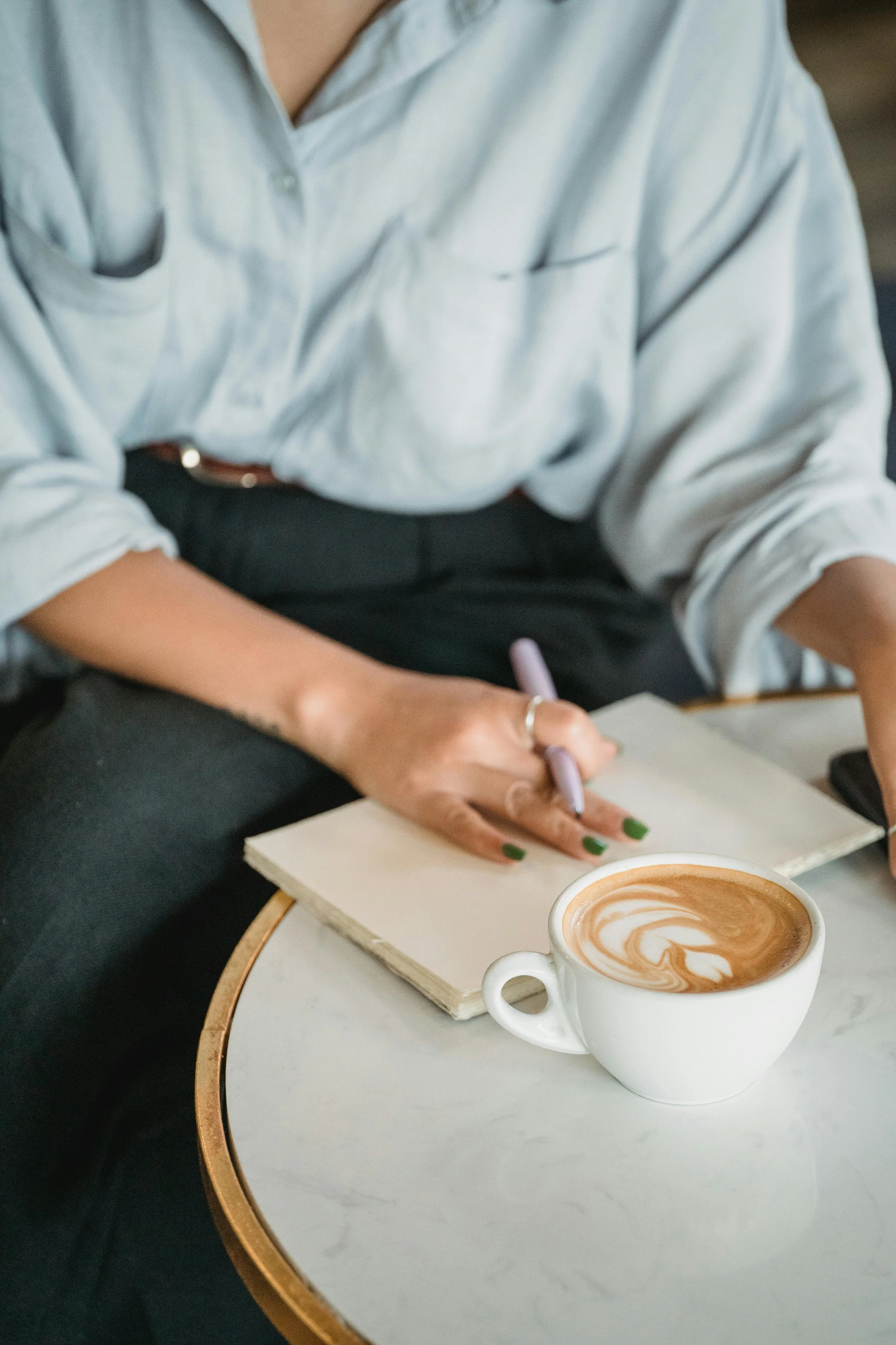 Person writing in a notebook with a latte on the table in a cafe.