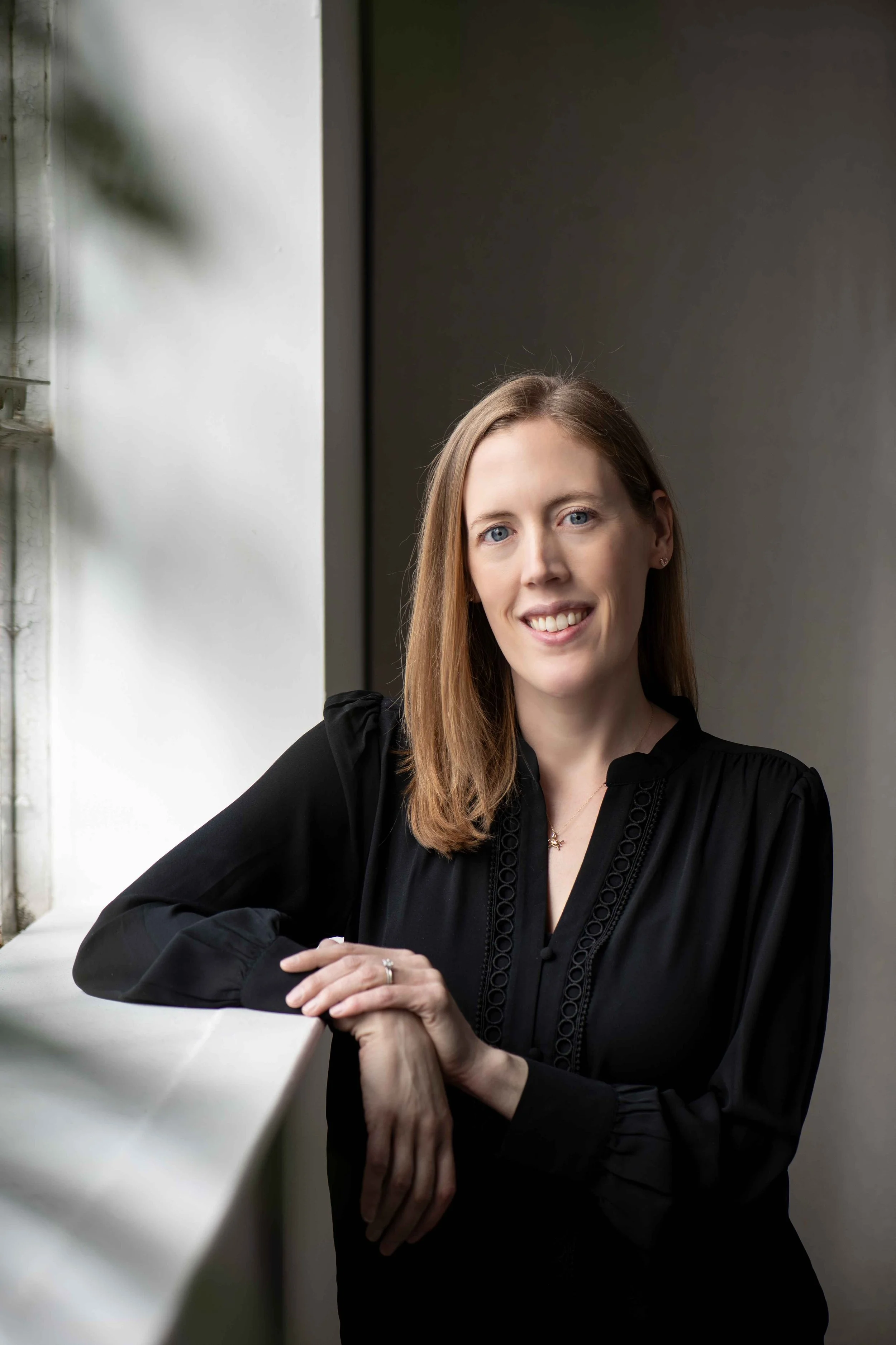 Dr. Christine Howard, a clinical psychologist who provides therapy and psychological testing, faces the camera and smiles while leaning on a white windowsill in an indoor setting with a gray wall.