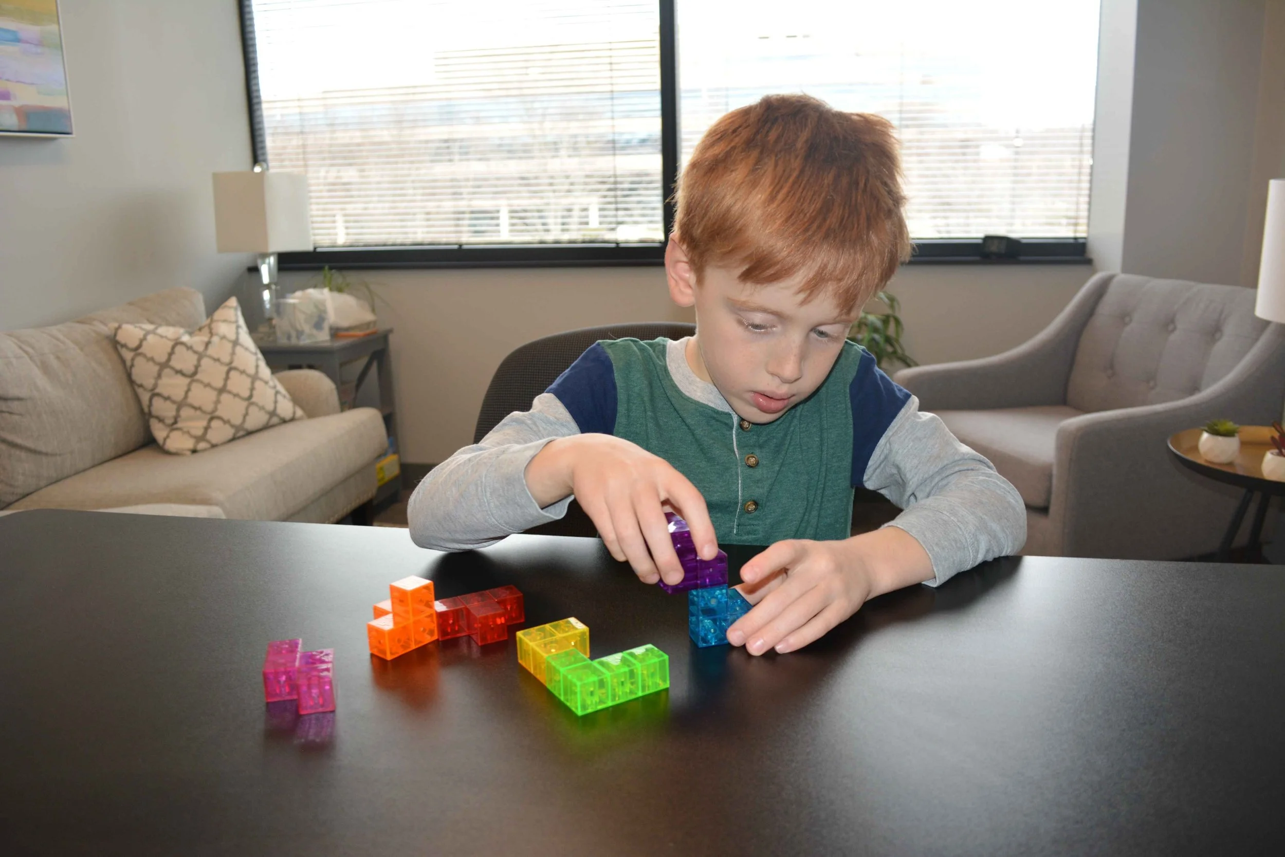 A 7-year-old boy with red hair playing with colorful transparent building blocks at a black table in Dr. Howard's office.
