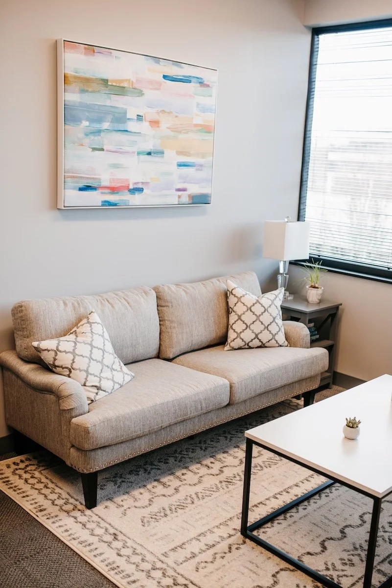 The inside of Dr. Christine Howard's therapy office. The room has a large window with horizontal blinds, a decorative rug, a gray carpeted floor, tufted chairs, and colorful artwork on the wall.
