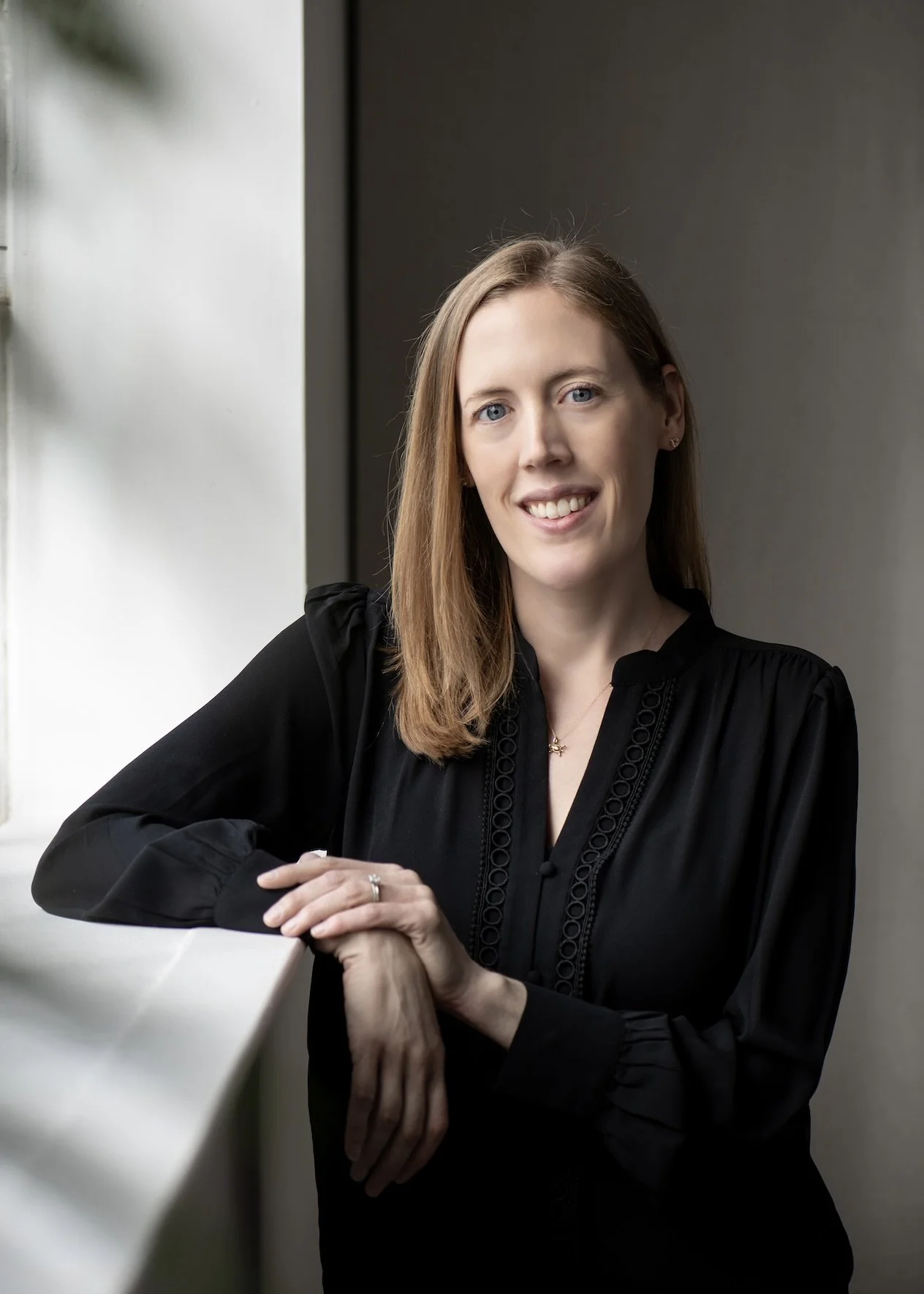 Dr. Christine Howard, a clinical psychologist who provides therapy and psychological testing, faces the camera and smiles while leaning on a white windowsill in an indoor setting with a gray wall.