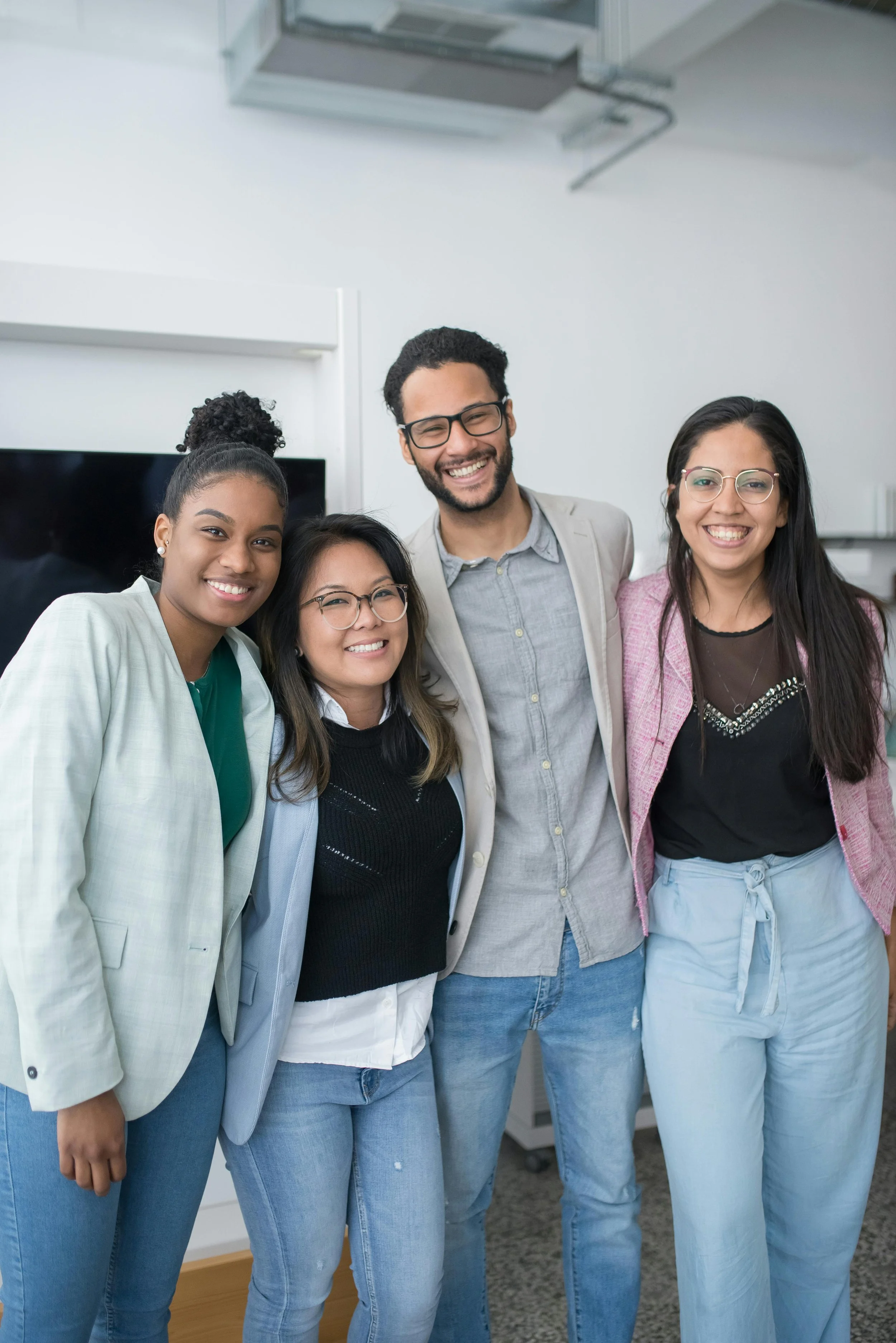 A diverse group of four young adults smiling and standing close together in an indoor setting, dressed in casual and business casual attire.