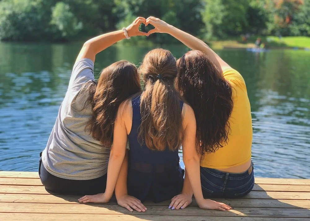 Three teenage girls sitting side-by-side on a wooden dock near a lake, with their backs to the camera, making a heart shape with their hands over the water, surrounded by green trees.
