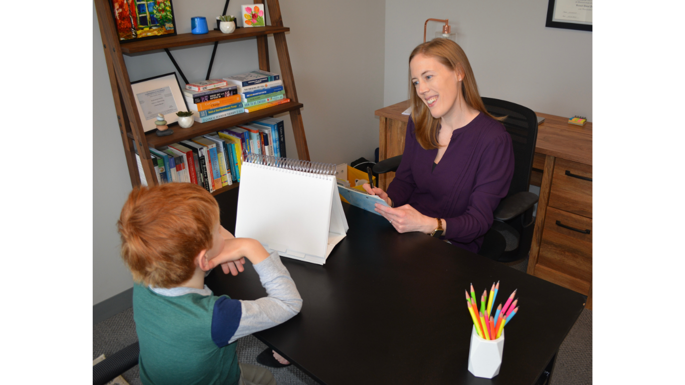 Dr. Christine Howard sits behind a black desk in her office, smiling and talking to a young boy with red hair, who is seated across from her. Dr. Howard and the boy work together to complete psychological testing.