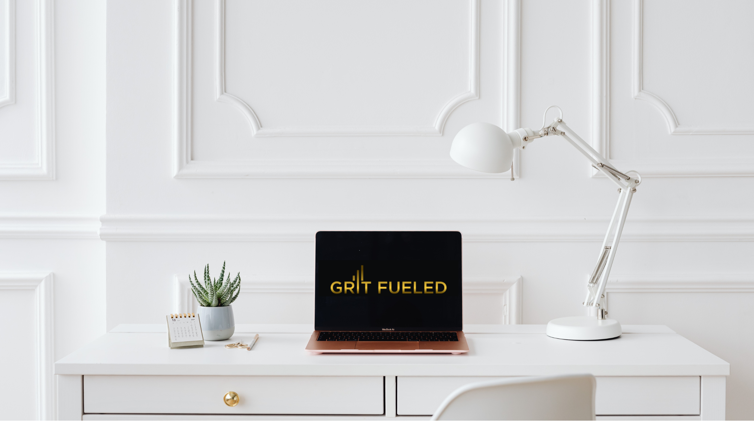 White desk with a laptop displaying 'Grit Fueled,' a potted plant, a small calendar, and a white desk lamp against a white panelled wall. Logo on the laptop reads, Grit Fueled.