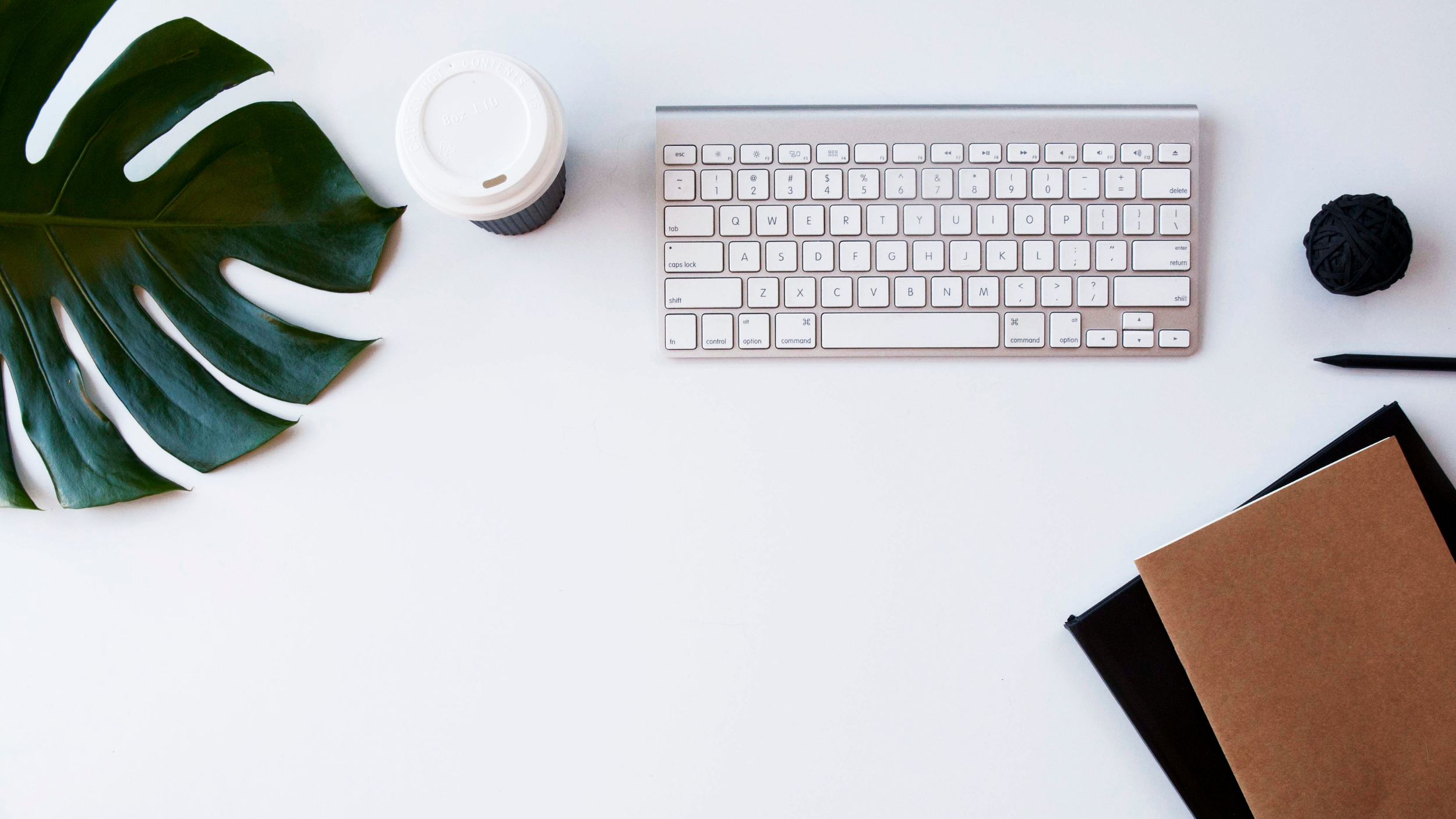 Minimalist entrepreneur's workspace with keyboard, coffee cup, leaf, notebooks, pen, and yarn ball on white desk.