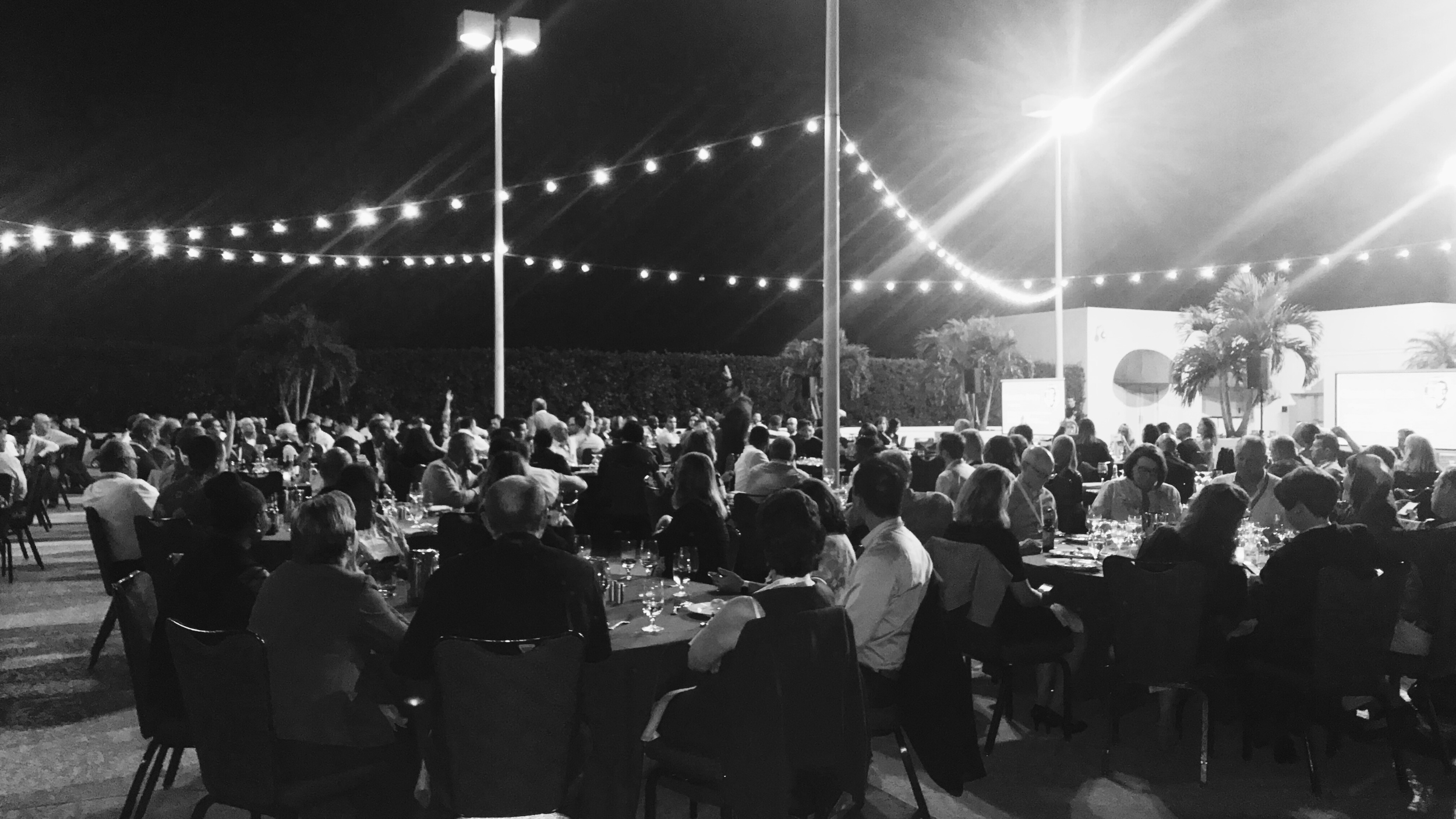 black and white photo of an evening business event, lit by string lights and the presenter captivating the audience in the middle of the tables