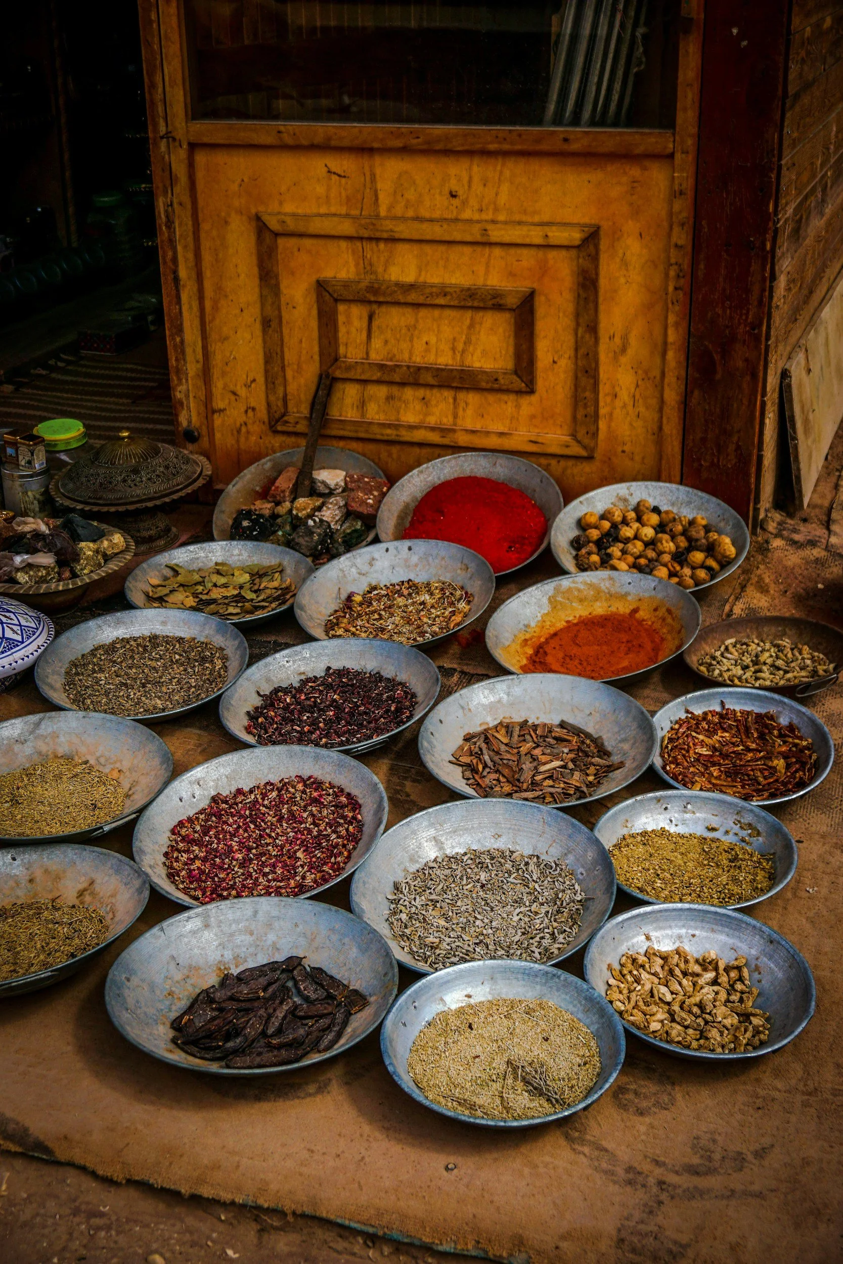 Assorted spices and herbs in metal bowls on a wooden floor, with a wooden door in the background.