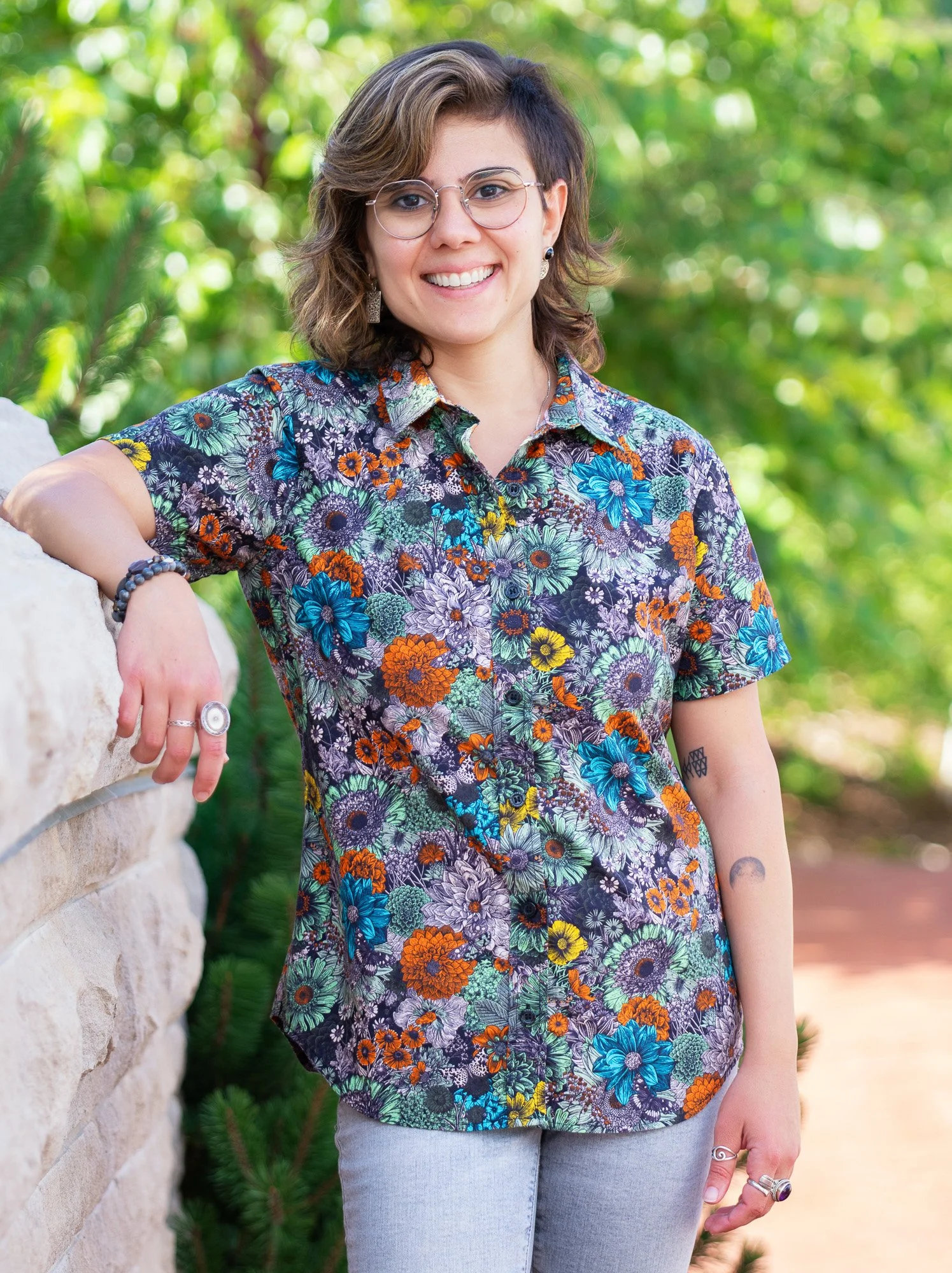A woman with glasses, short wavy brown hair, wearing a colorful floral shirt, standing outdoors against a stone wall and green foliage, smiling.