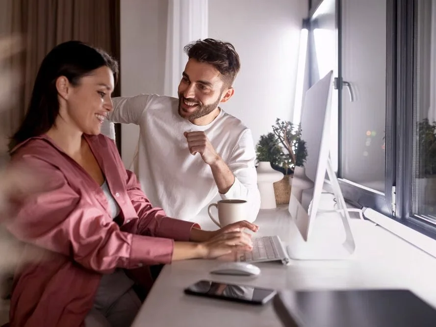 Man and woman smiling while sitting at a computer desk and drinking coffee