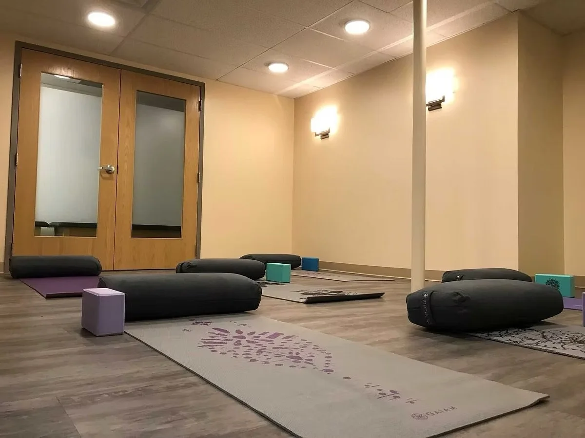 Yoga studio with black bolsters, yoga mats, foam blocks, and decorative rugs on a wooden floor, with beige walls, double glass doors, and soft wall lighting.