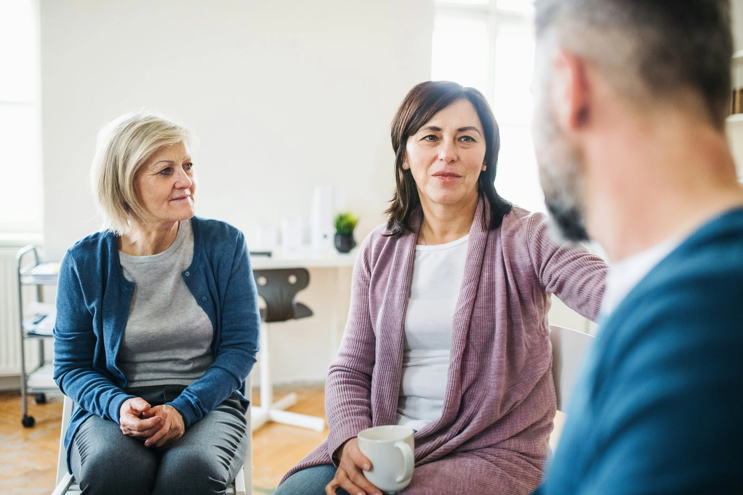 Three women sitting in a circle during a conversation, in a bright room with windows and a small table in the background.