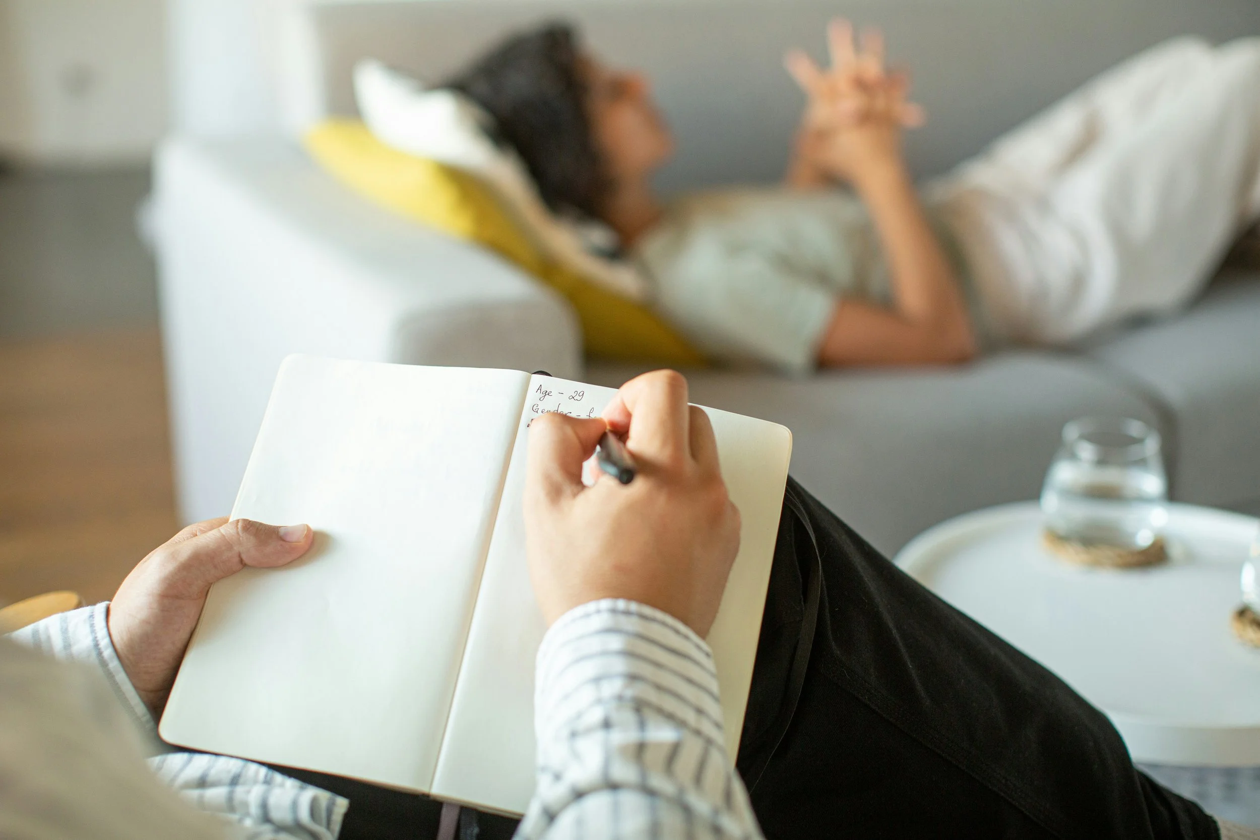 Person sitting on a couch writing in a notebook while another person lies on a couch in the background.