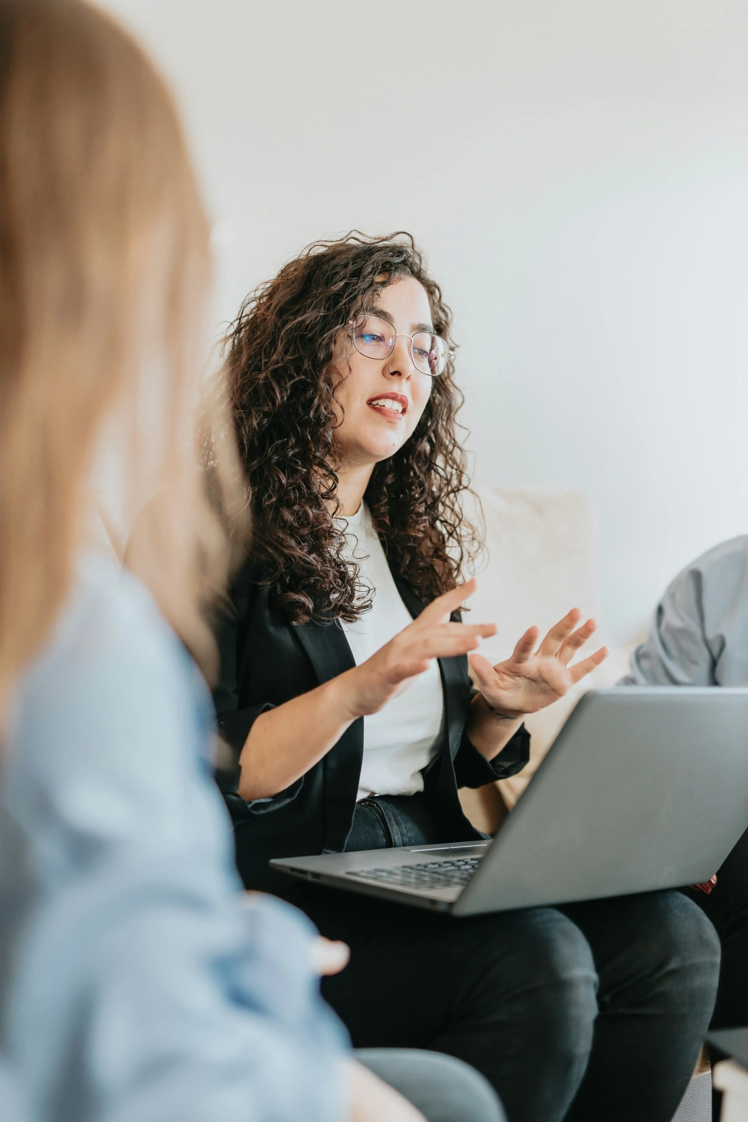 Woman with curly hair speaks while gesturing, sitting with a laptop on her lap in a group discussion with two others.