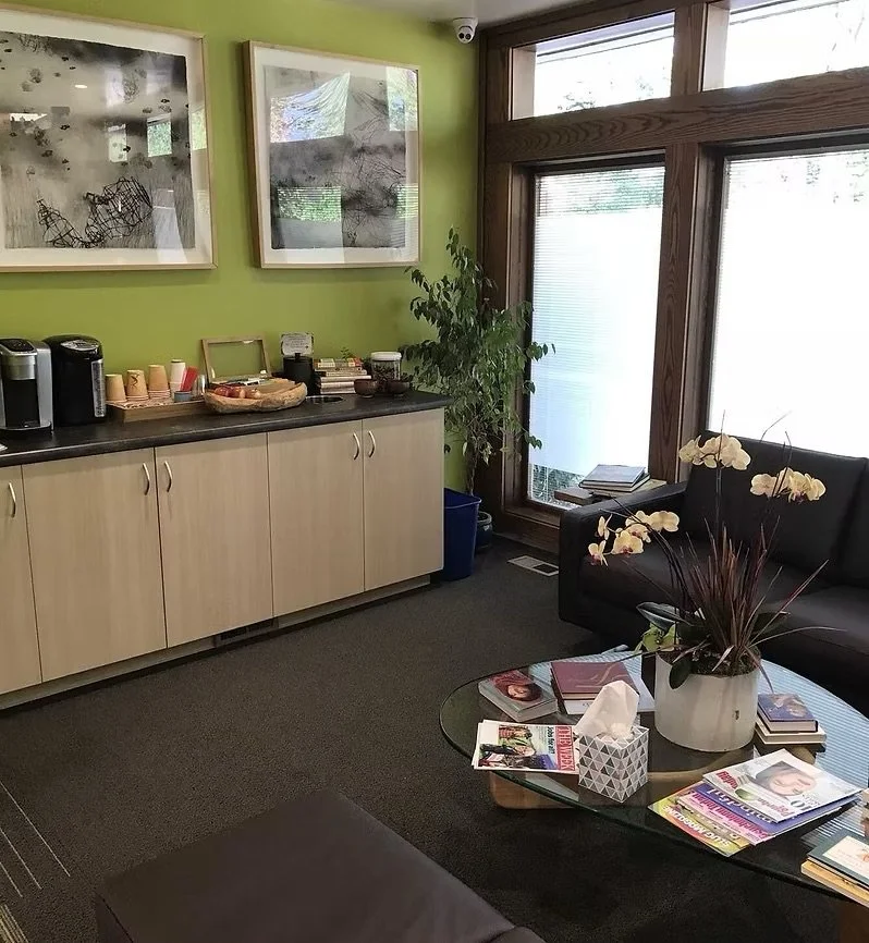 Office lounge with a black sofa, glass coffee table with magazines and tissue box, green wall with artwork, coffee station with cups and coffee makers, large window with wooden frame, potted plant near the window.
