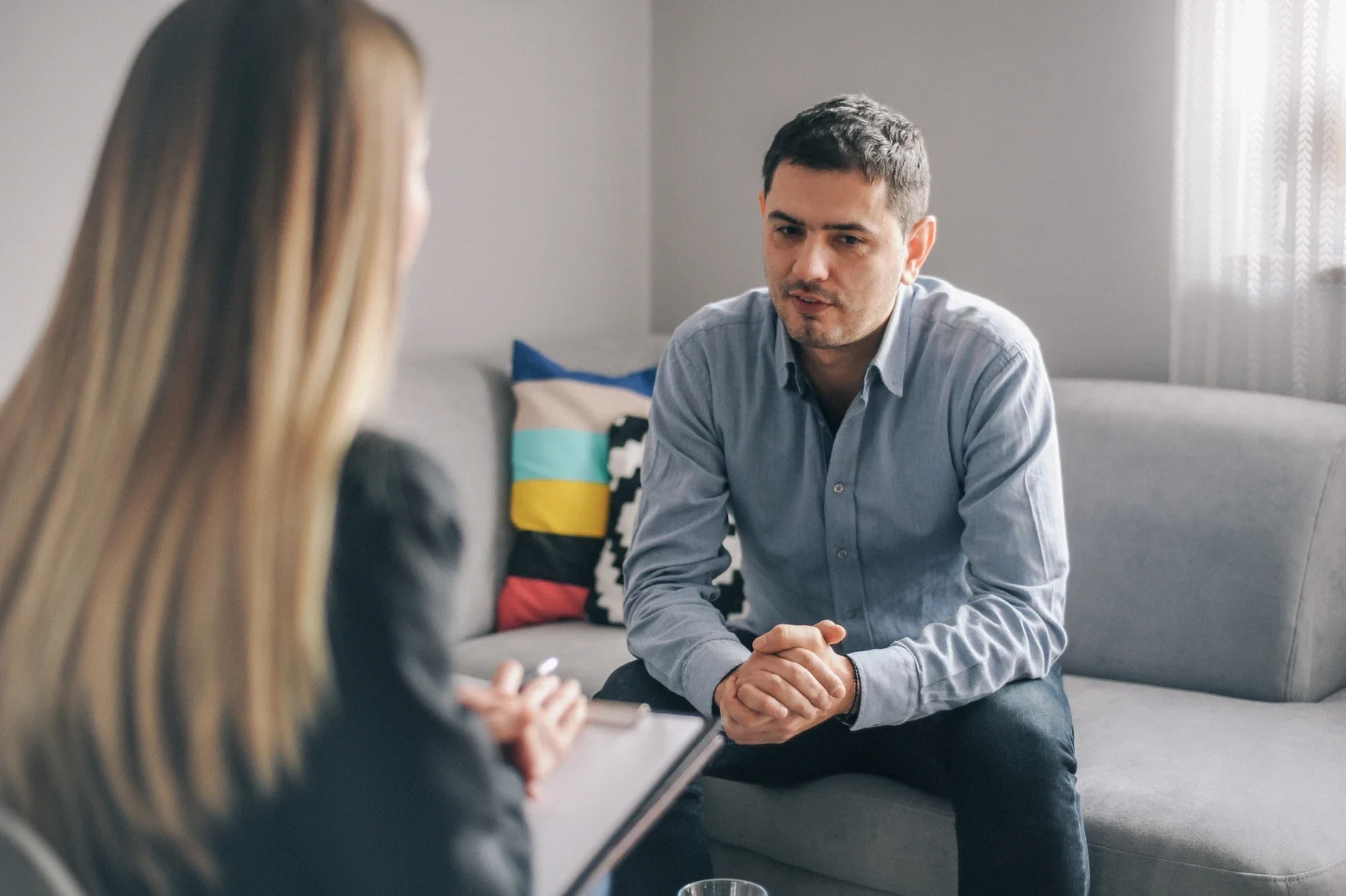 Male patient talking to female therapist in a room with gray pain and a gray couch. The male looks anxious.