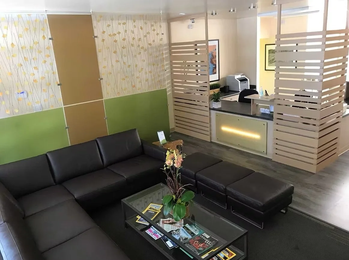 Modern hotel lobby with black leather sectional sofa, glass coffee table with magazines, decorative plant, reception desk with computer, and wooden partition panels.