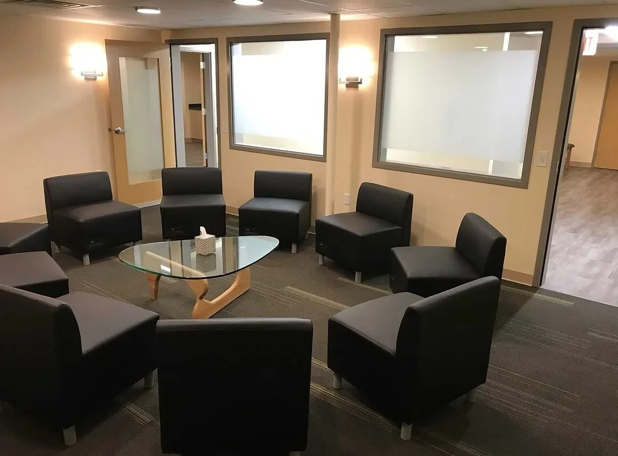 An empty waiting room with eight black chairs arranged in a semi-circle around a glass coffee table, with a tissue box on top, beige walls, wall-mounted lights, and windows with frosted decals.