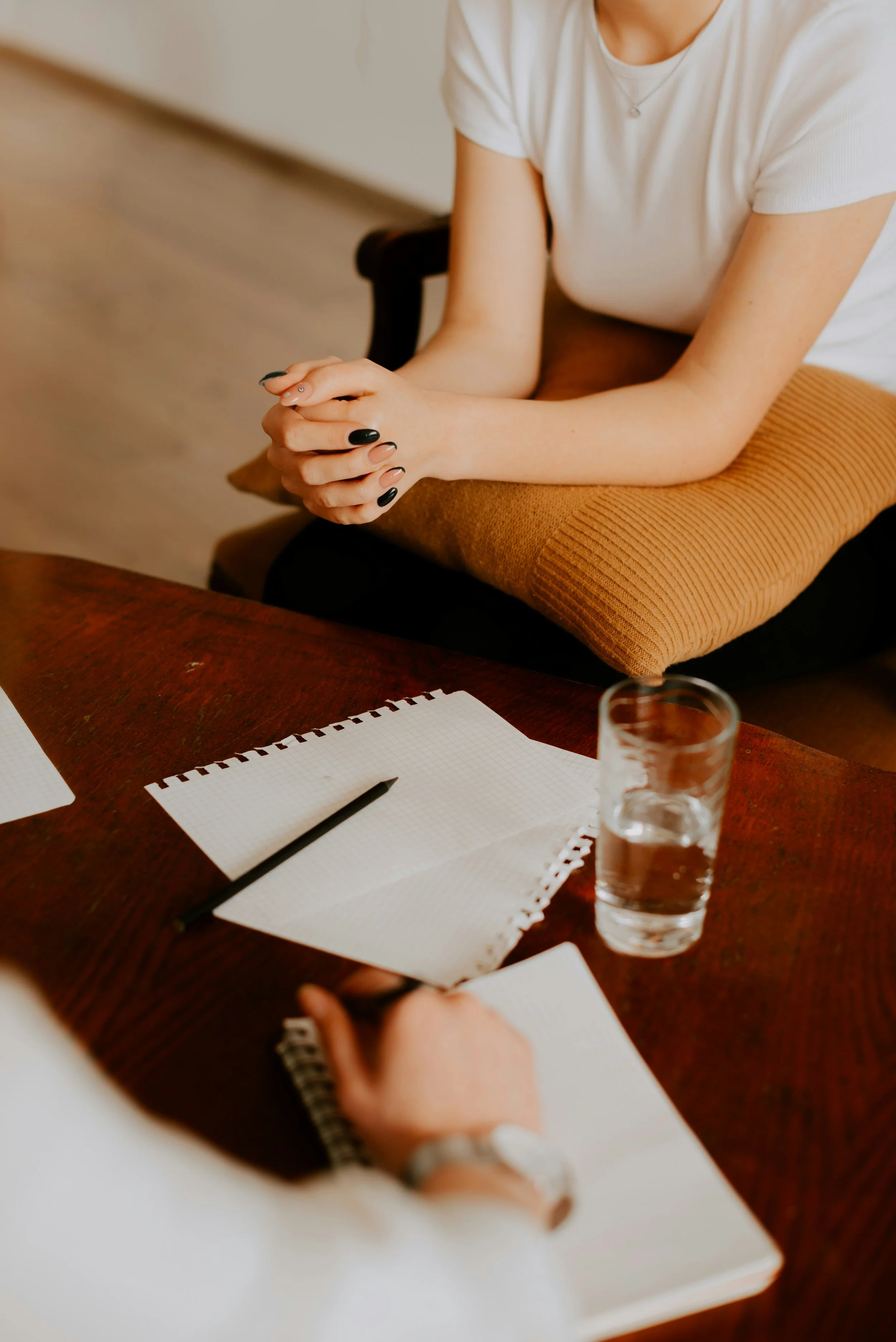 A woman wearing a white shirt sitting at a table with a notebook, pen, glass of water, and another person's hand holding a notepad in front of her.