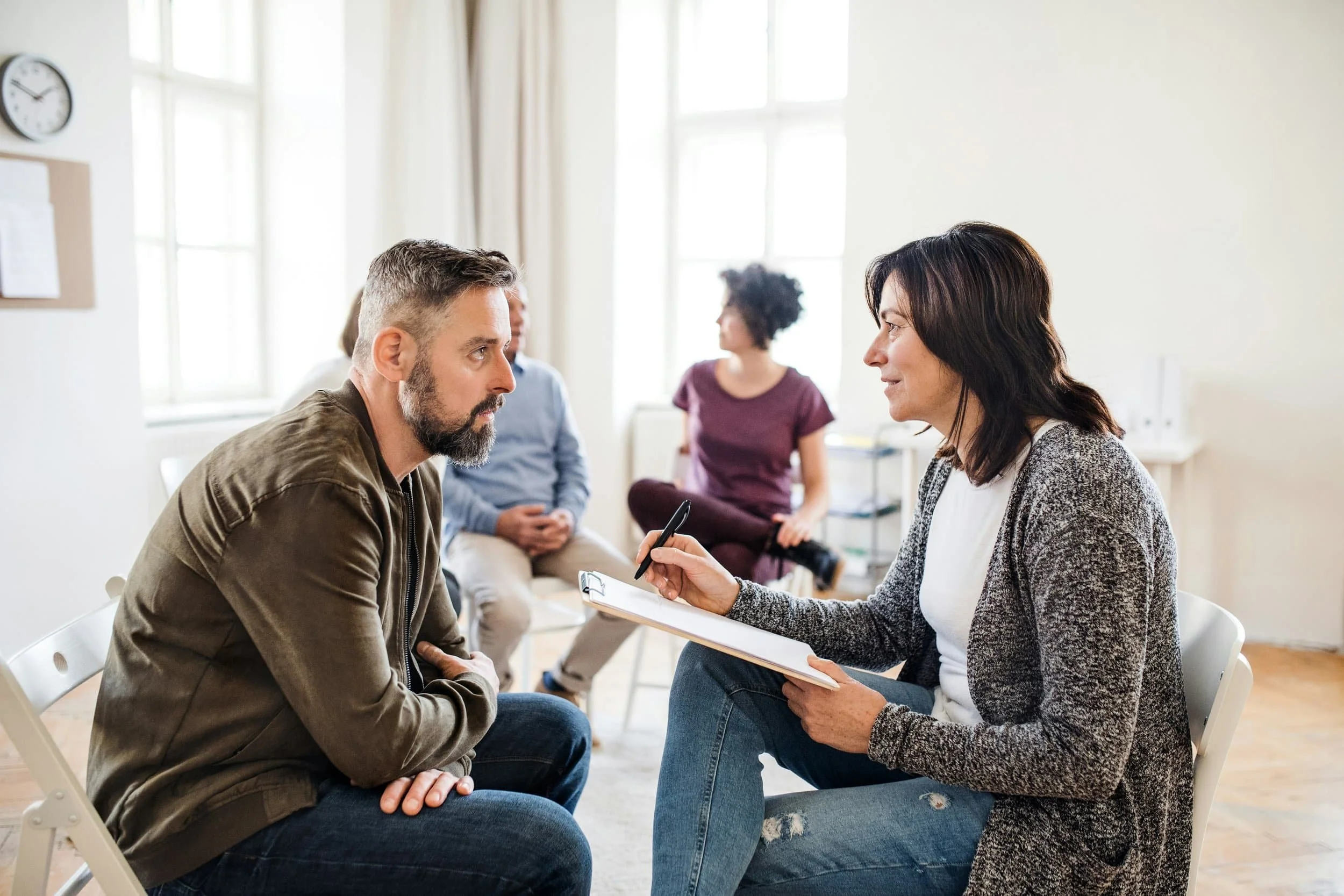 A therapist or counselor is speaking with a man during therapy session, with other clients in the background in a well-lit office.