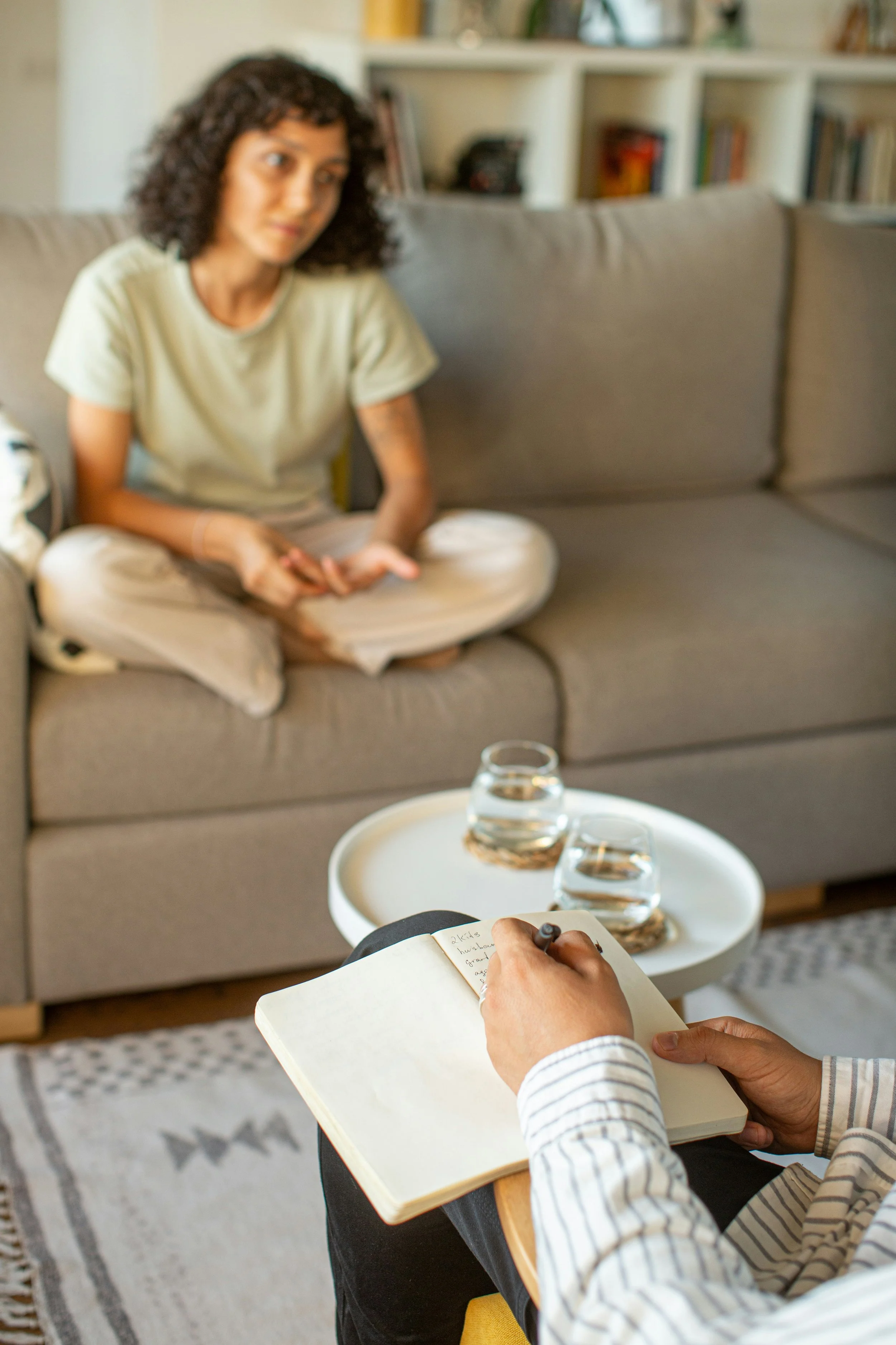 A woman sitting on a gray couch during a therapy session, talking with a therapist who is taking notes. The therapist has a notebook and a pen, with two glasses of water on a small white table in front of them. The room has a bookshelf in the background.