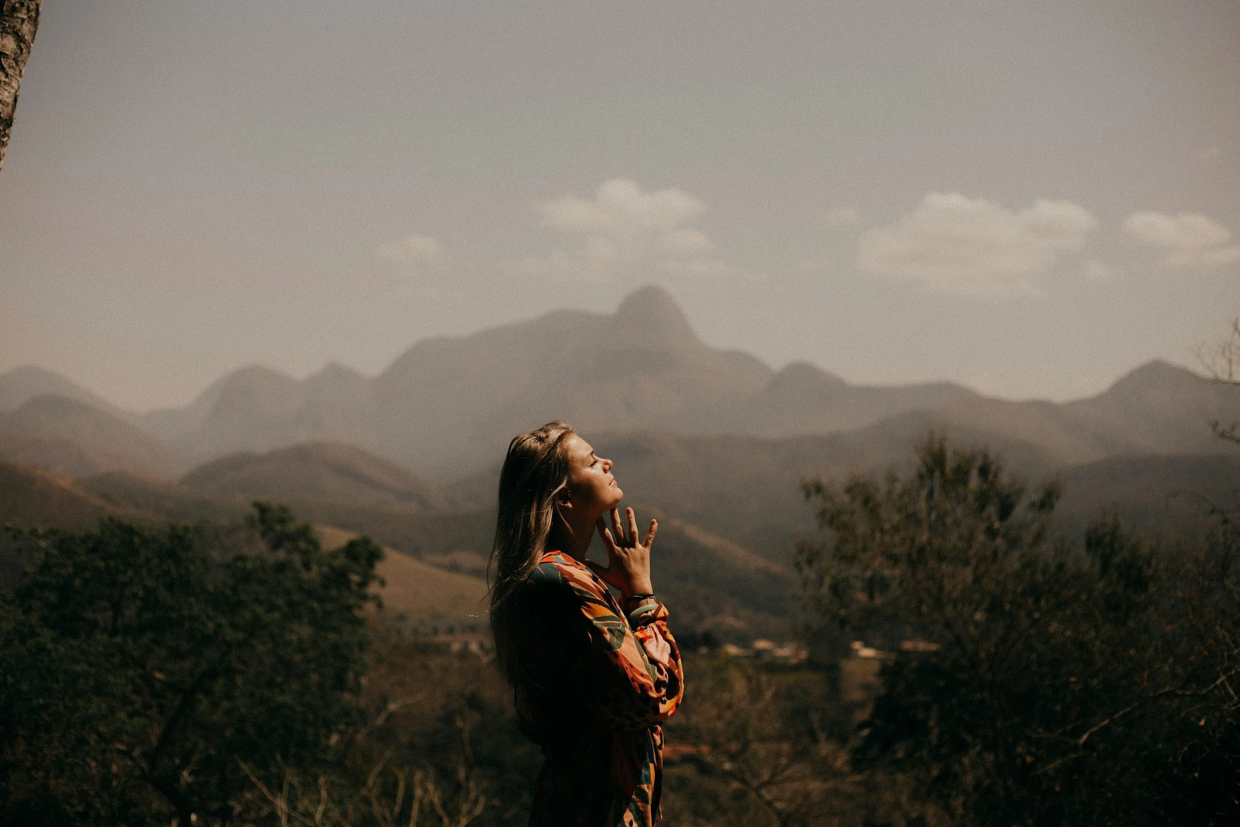 A woman standing outdoors with mountains in the background, eyes closed, enjoying the scenery.