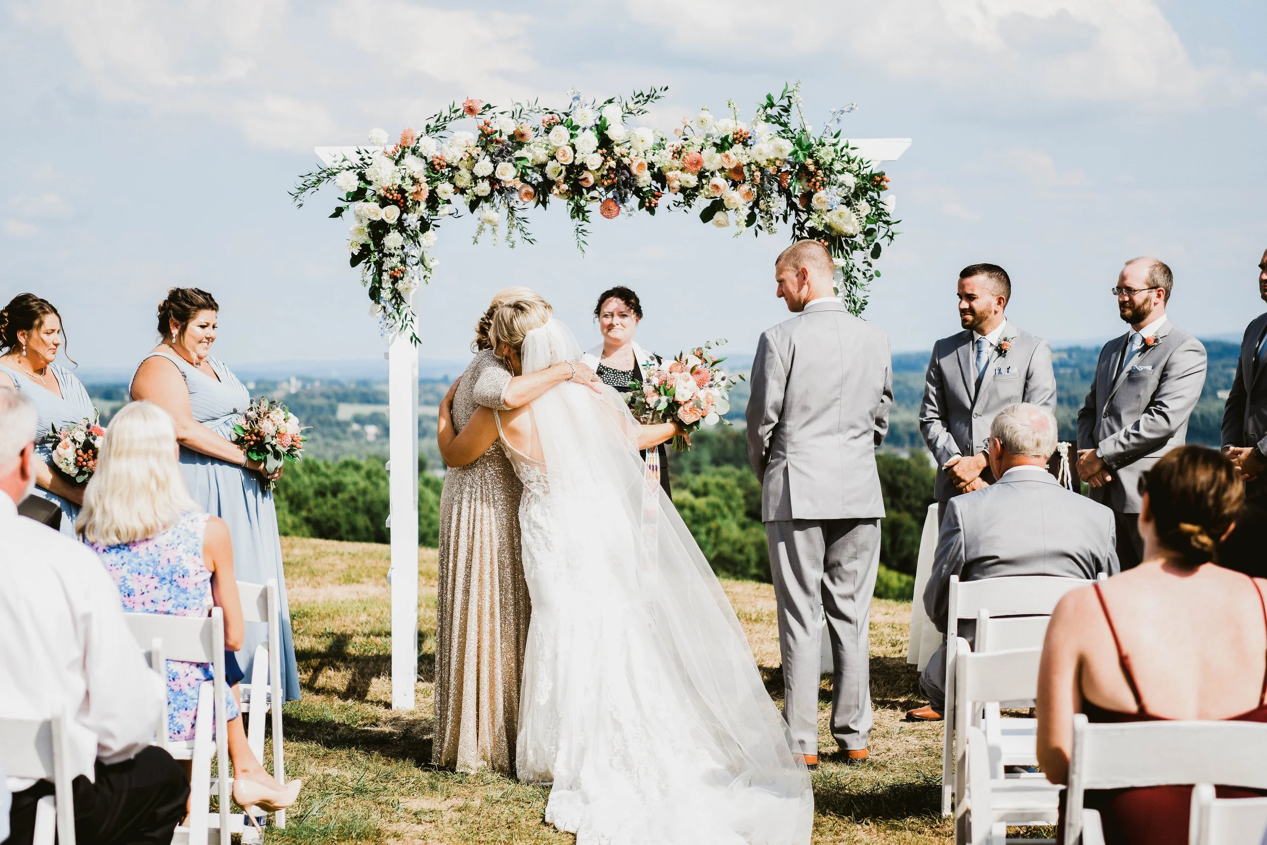 An outdoor wedding ceremony with a bride and groom in front of a floral arch, surrounded by bridesmaids and groomsmen. Attendees are seated on a grassy hillside with scenic views.