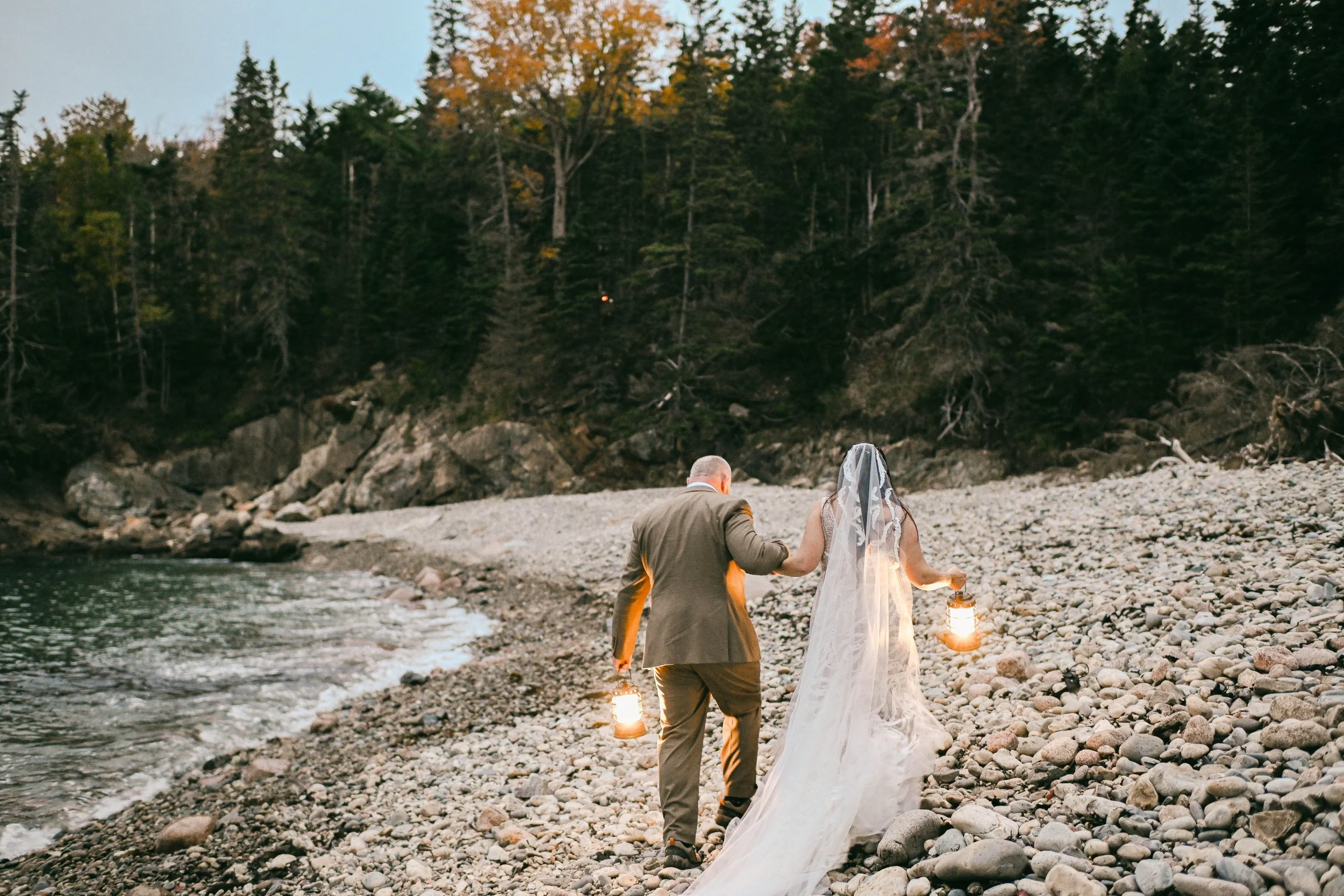 A couple dressed in wedding attire walking on a rocky beach holding lanterns, with a forested cliff in the background during dusk or dawn.