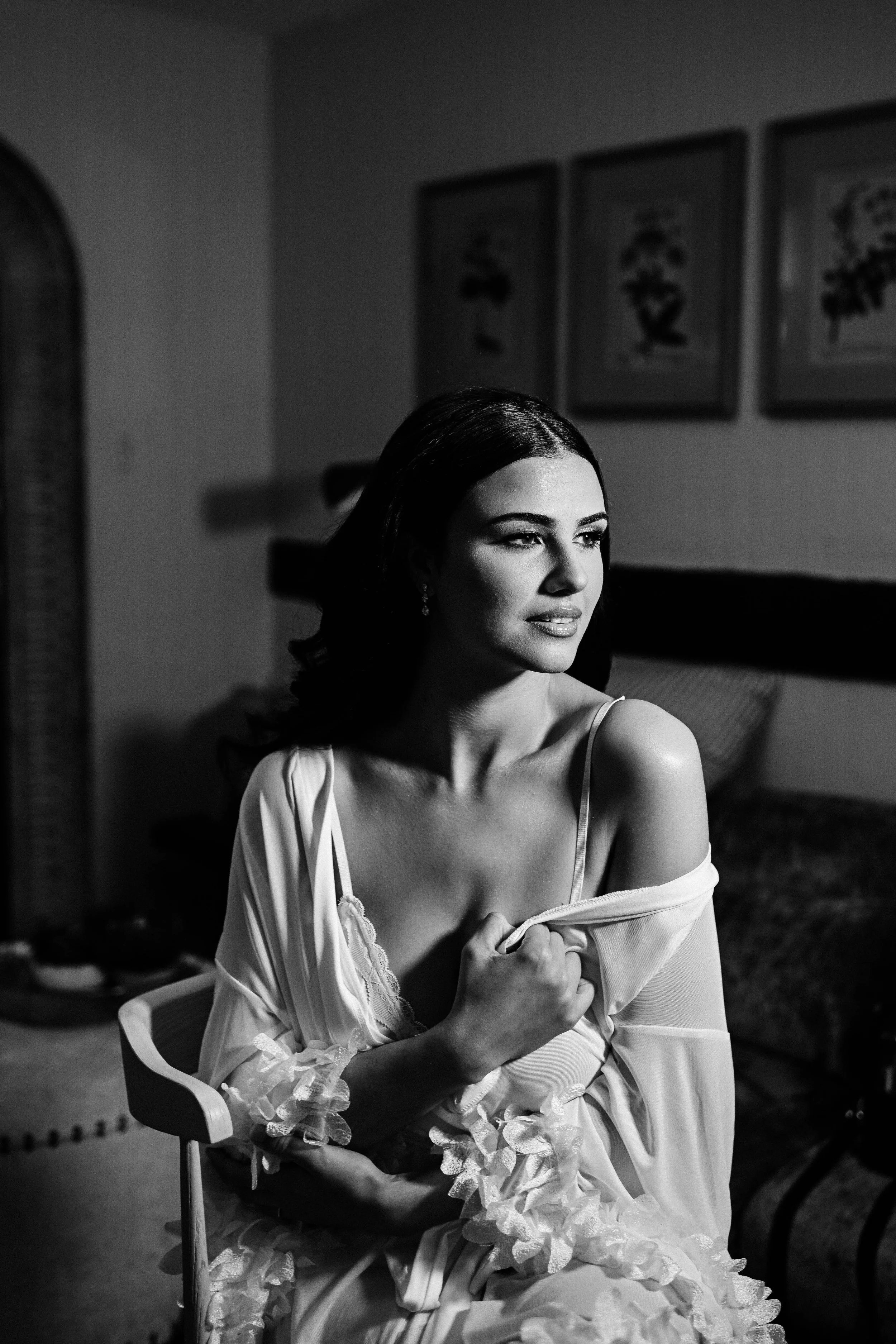 A woman with dark hair in a robe, sitting in a room with three framed pictures on the wall, looking away thoughtfully in a black and white photo.