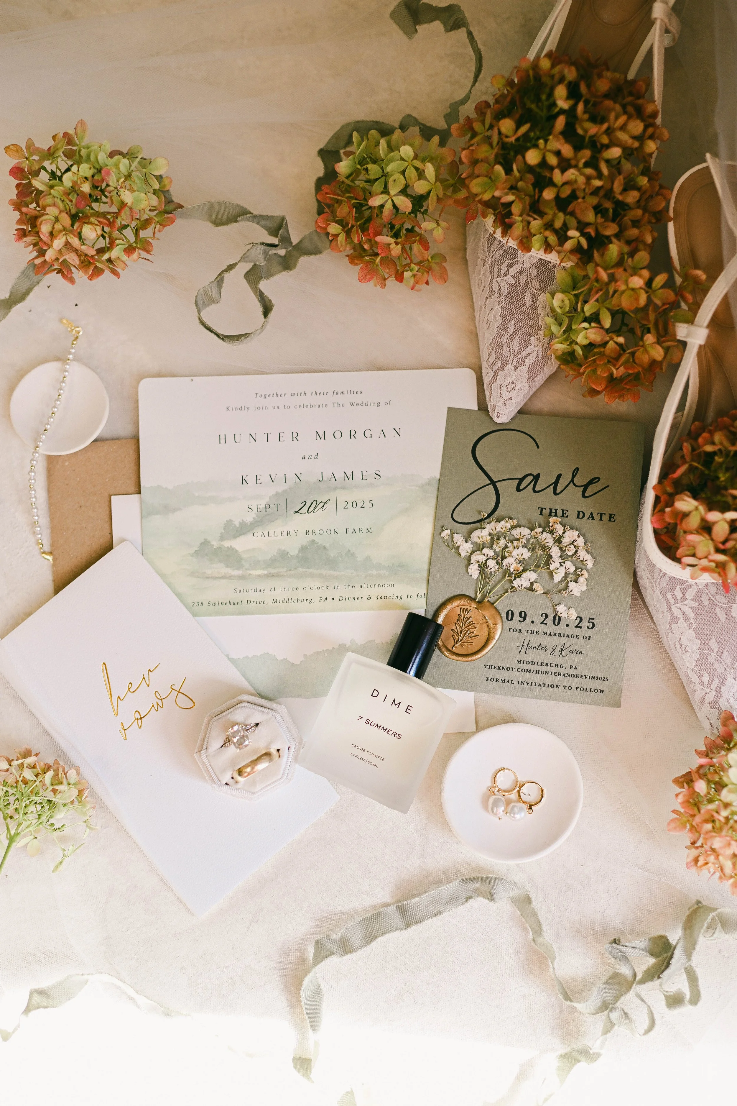 Wedding save-the-date and invitation cards, rings, perfume, jewelry, and floral arrangements on a table.