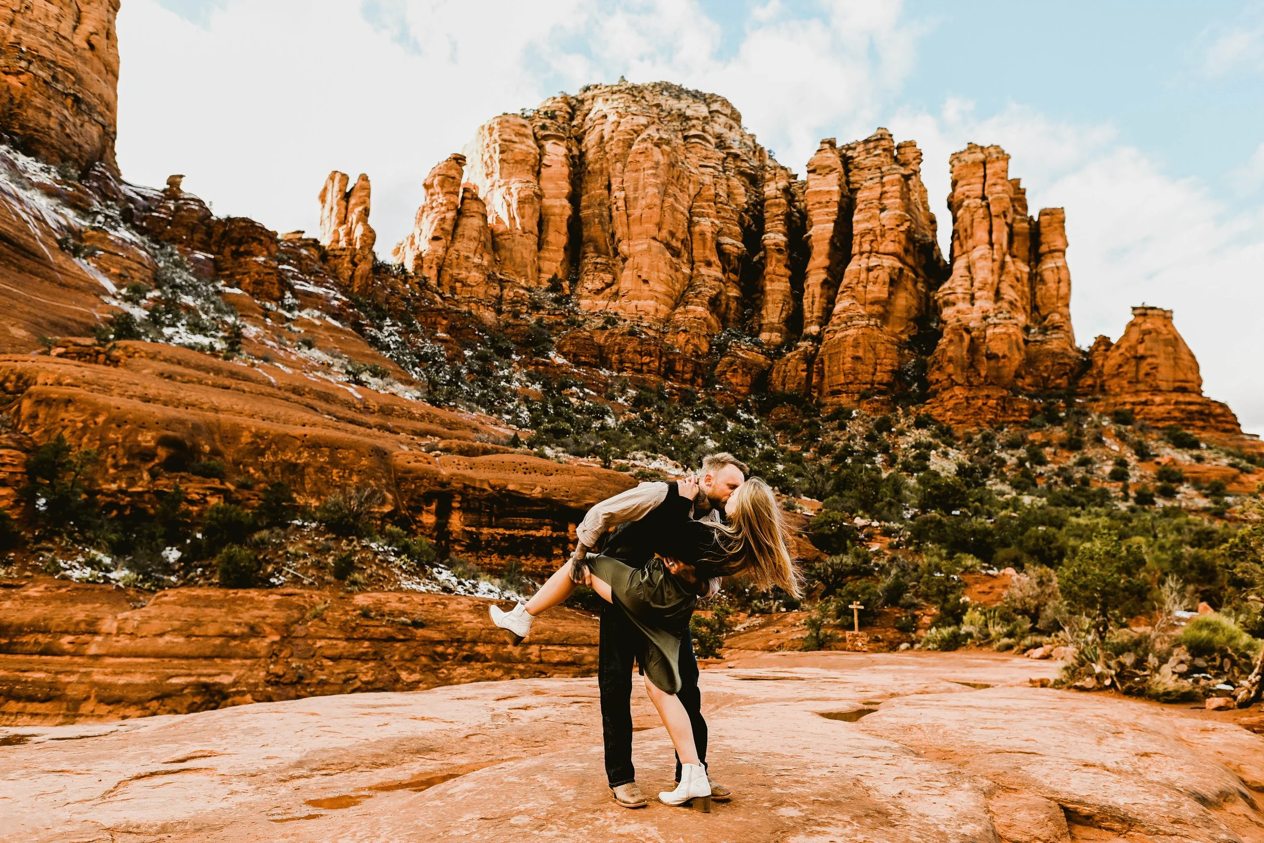 A couple is kissing in a desert landscape with red rock formations and sparse vegetation, one person is lifting the other.
