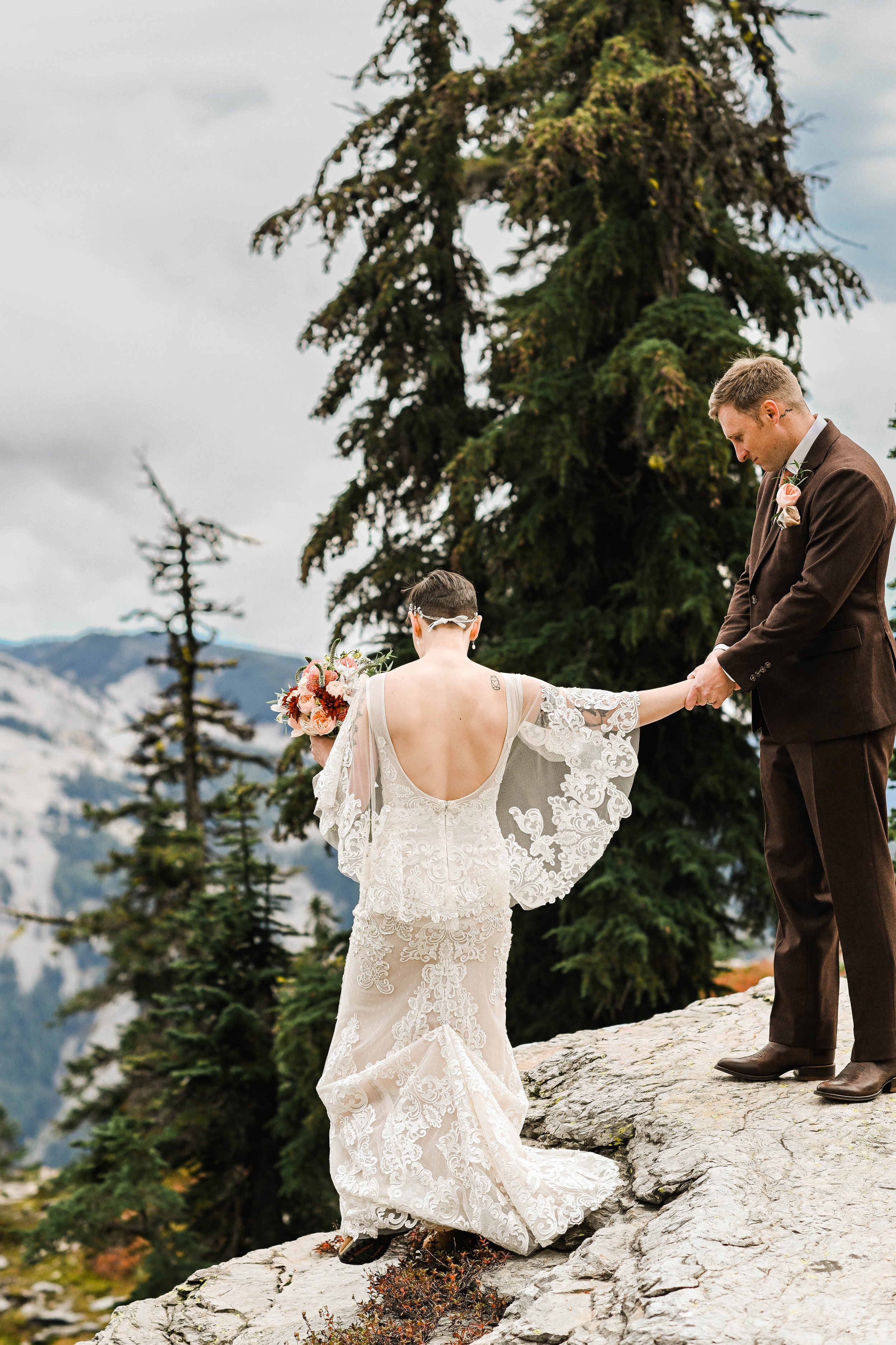 A bride and groom holding hands on a rocky ledge in a mountain setting. The bride wears a lace wedding dress with an open back and holds a bouquet of flowers. The groom is dressed in a brown suit with a boutonniere. Tall pine trees and mountain peaks