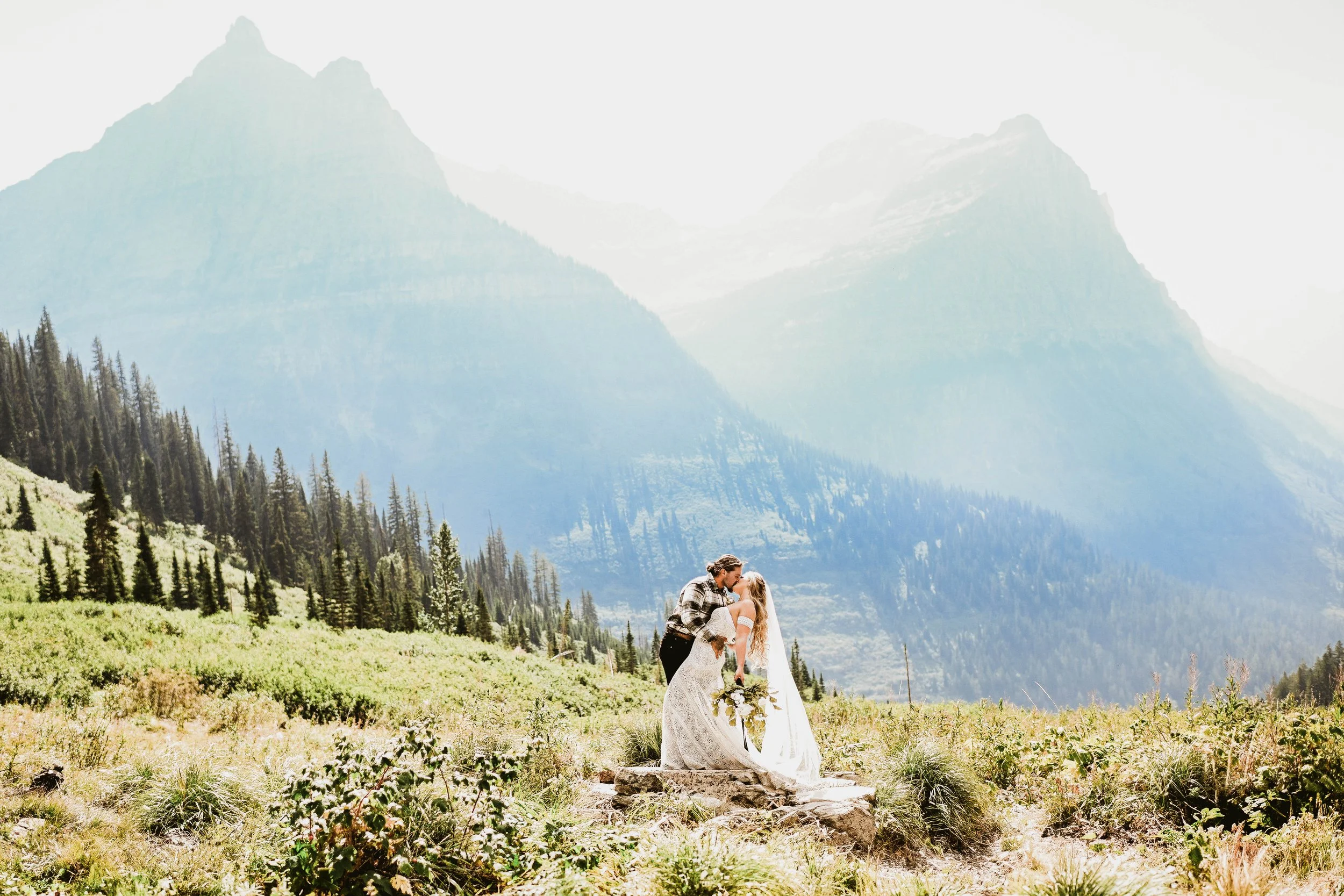 A couple in wedding attire embracing and kissing in a mountain meadow, with forested slopes and towering mountains in the background.