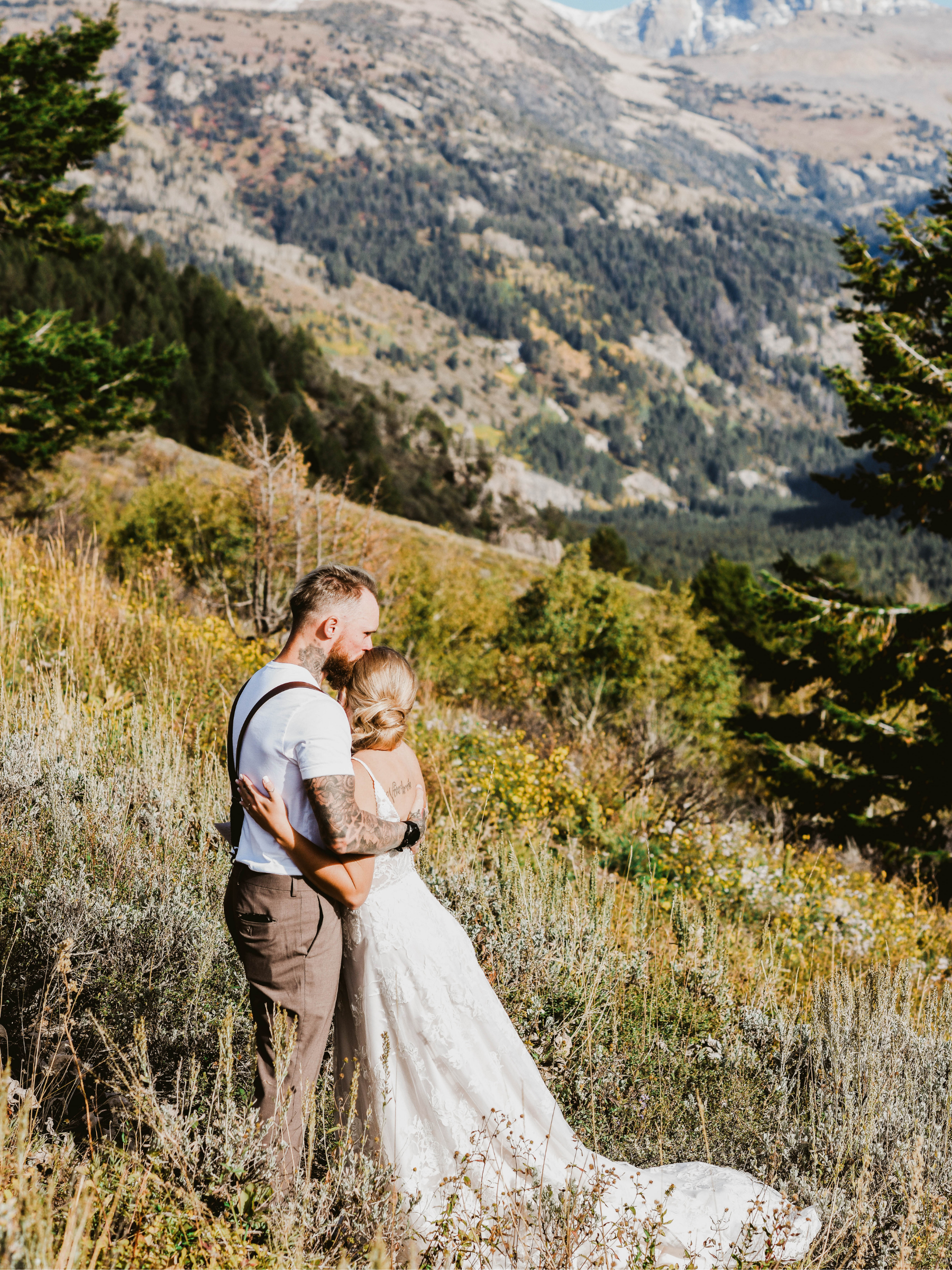 A couple in wedding attire embracing in a scenic mountainous outdoor setting with trees and rolling hills in the background.