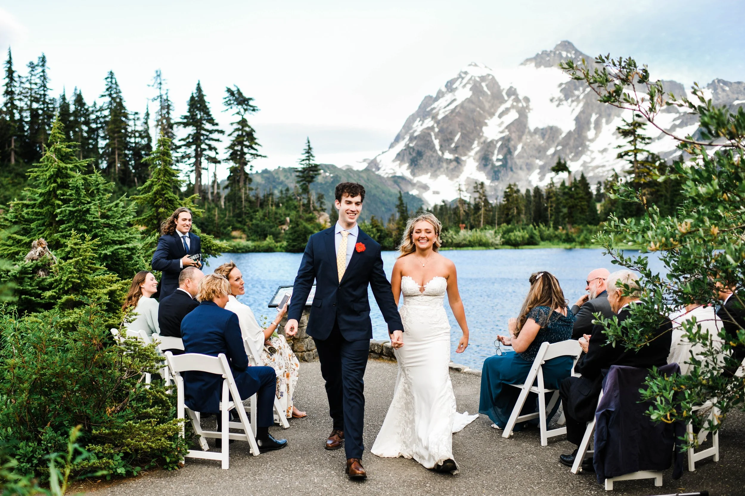 A bride and groom walking hand in hand during their outdoor wedding ceremony by a lake with mountains and trees in the background.