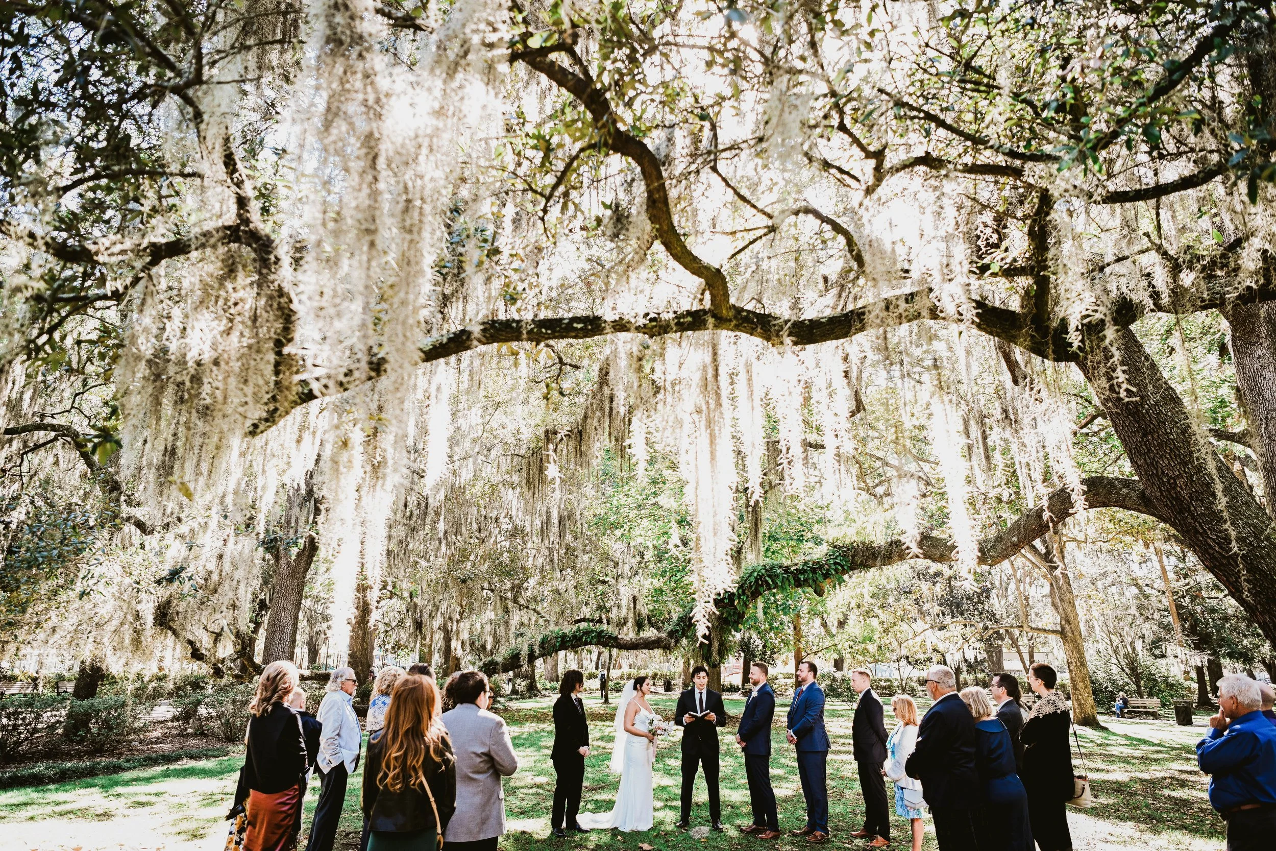 A wedding ceremony taking place outdoors under large, hanging white blossoms on trees with a small group of people gathered around a bride and groom.