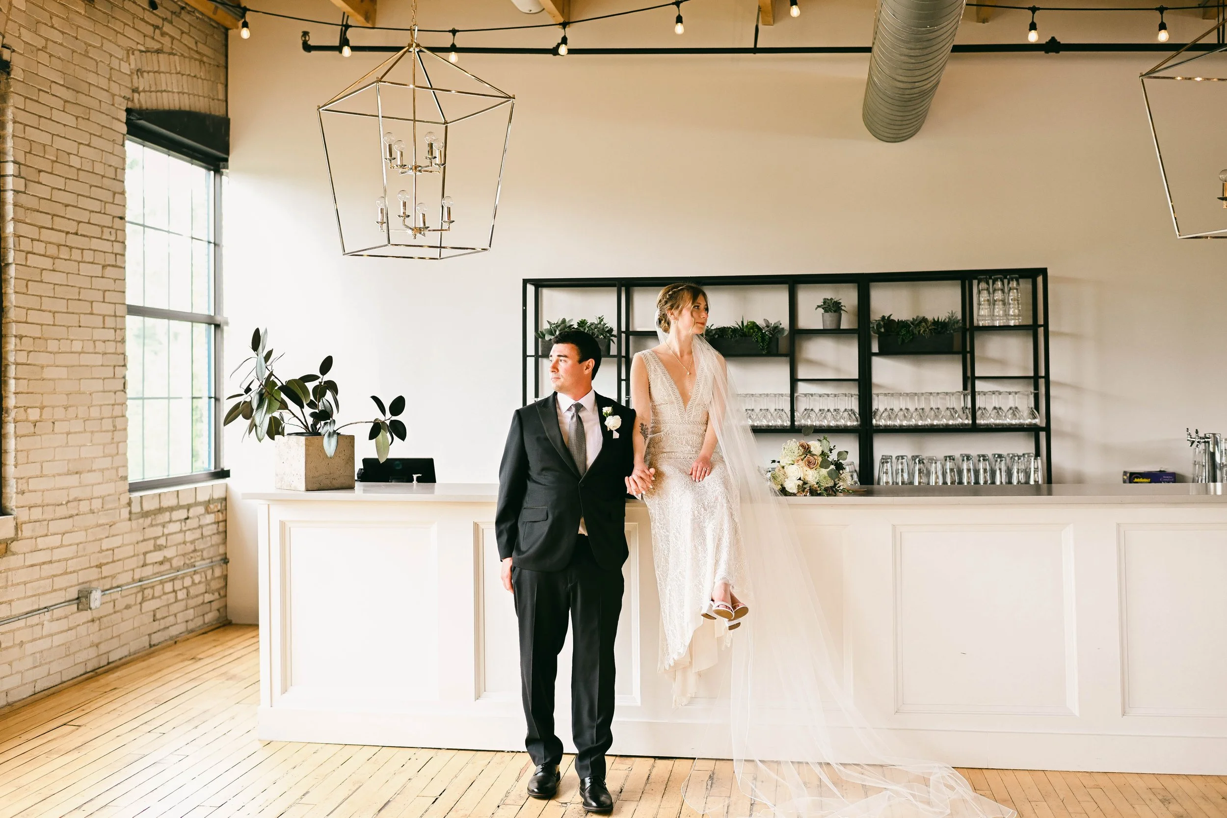 A bride and groom standing at a bar in a wedding venue. The bride is sitting on the bar, wearing a white wedding dress and veil, holding a bouquet, and the groom stands beside her in a black suit and tie. The venue has brick walls, large windows, and