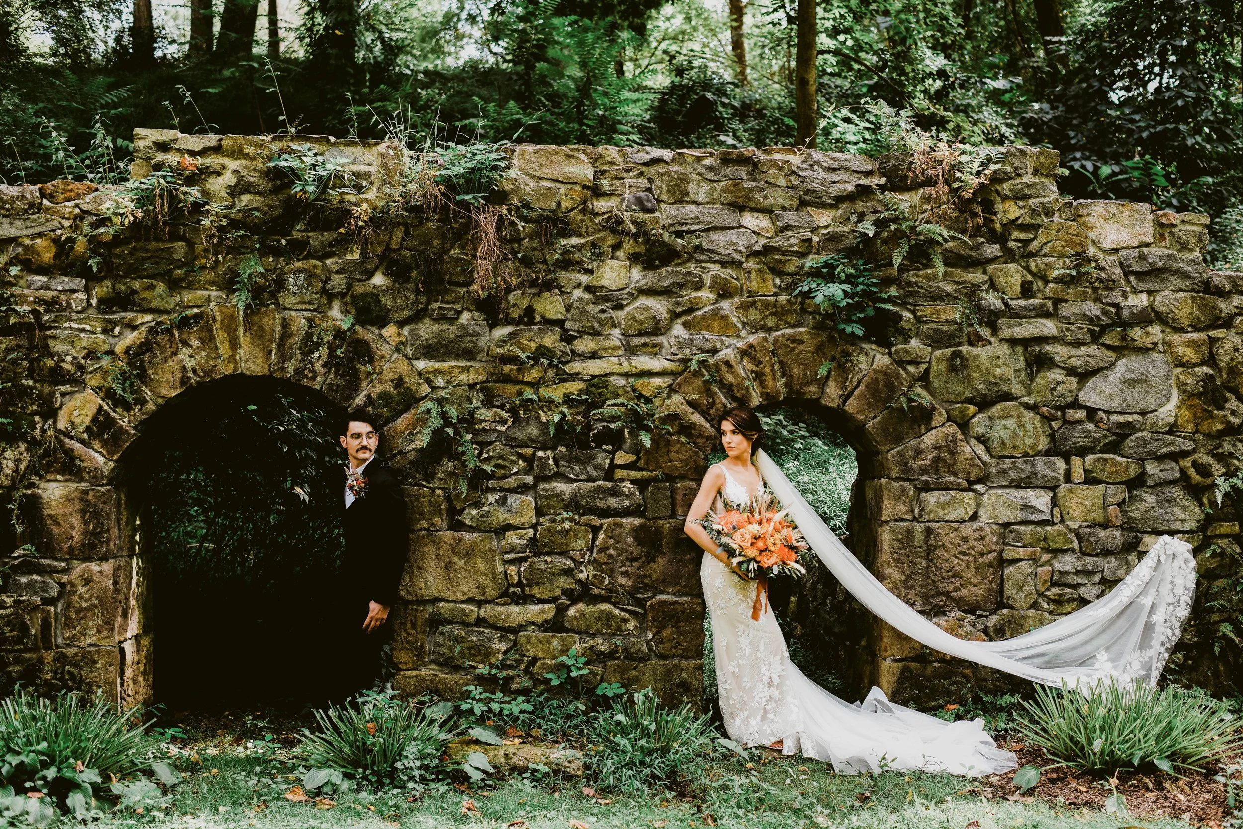 Bride in a white wedding dress holding a bouquet, standing in front of an old stone wall with two arches, one with a man in a dark suit and tie partially visible in the left arch, the other with lush greenery in the background.