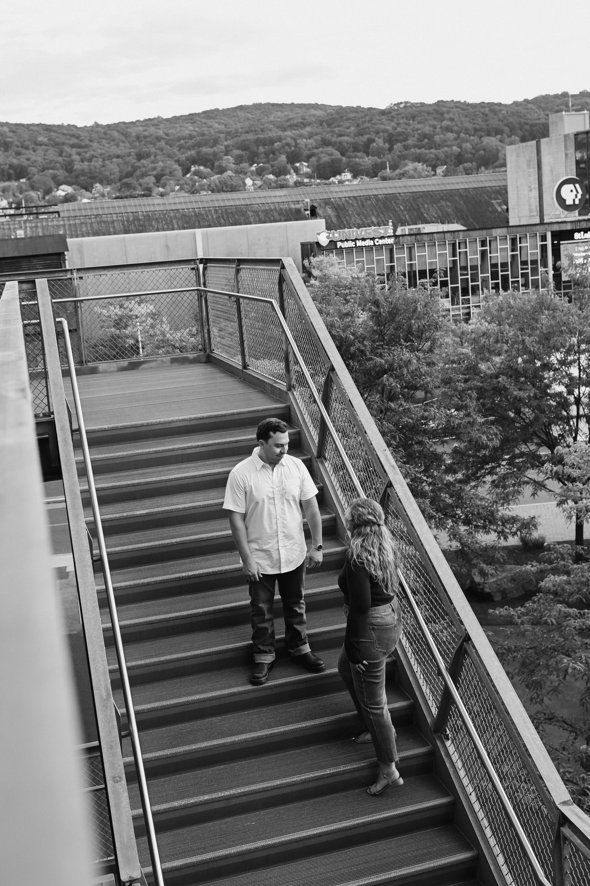 Two people, a man and a woman, are standing on an outdoor staircase and engaging in conversation. The man is wearing a light-colored short-sleeve shirt and dark pants, while the woman has long curly hair and is wearing a dark top and light pants. In 